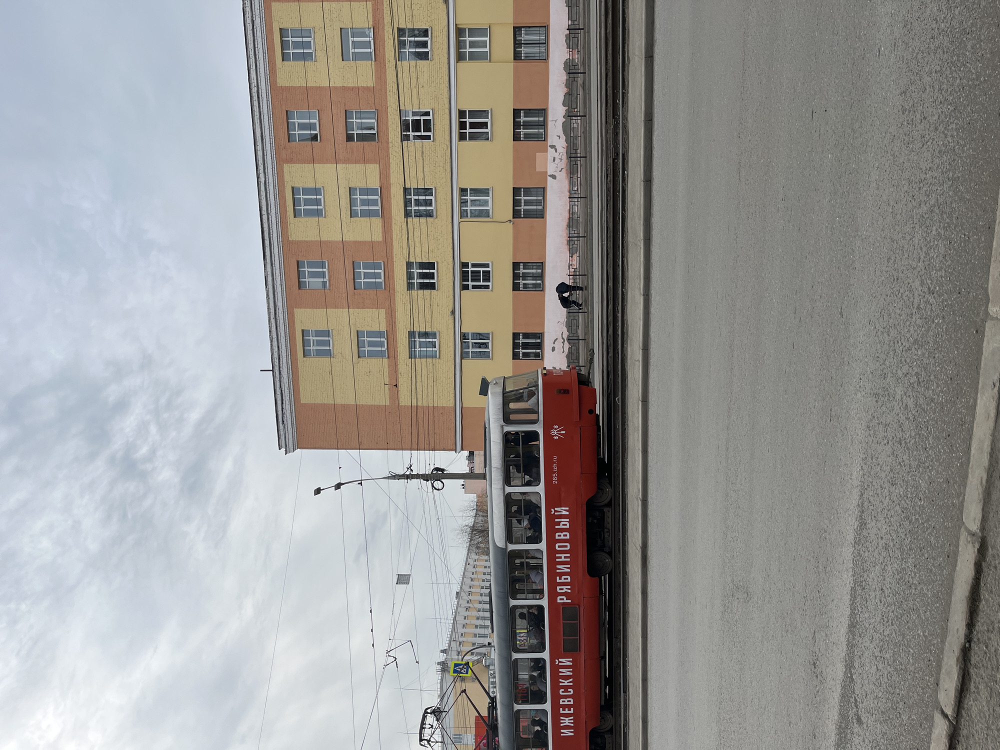 The image depicts a street scene in Izhevsk, Russia, featuring a red and white tram traveling along tram tracks parallel to a gray asphalt road. The side of the tram displays the Cyrillic inscription "ИЖЕВСКИЙ РЯБИНОВЫЙ" (Izhevsk Rowan). A multi-story building with alternating beige and peach-colored facade panels and evenly spaced windows is visible adjacent to the tracks. A person in dark clothing is near the tram tracks. Overhead, electrical wires and poles support the tram's power system. The sky is overcast with light gray clouds. The image is rotated 90 degrees counterclockwise.