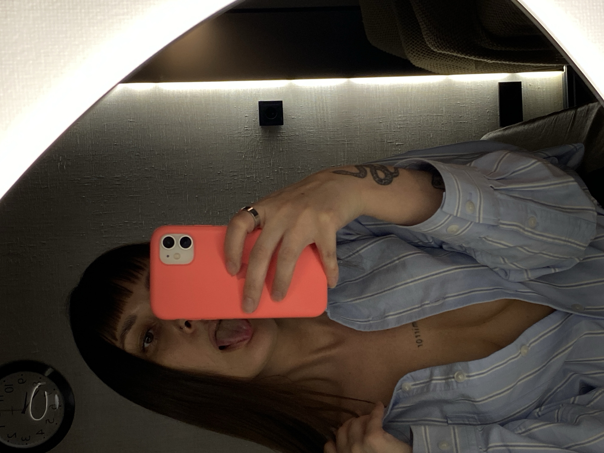 A person is taking a selfie in a mirror with a coral-colored iPhone. The person has dark hair with bangs, and is sticking their tongue out. The person is wearing a light blue and white striped button-down shirt, with the top few buttons undone. There is a snake tattoo on their left arm and a tattoo on their chest. The mirror is lit by lights above and to the side. A clock is visible in the lower left corner of the mirror. The location is unknown, and the image does not provide information about Votkinsk, Russia.