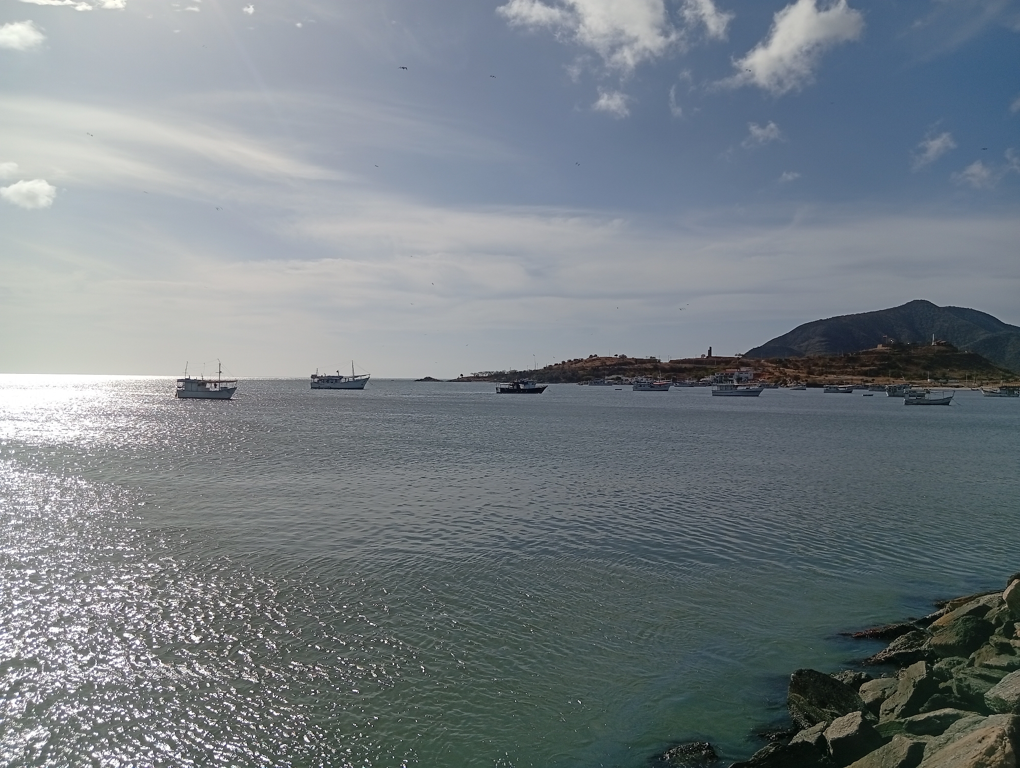 The image depicts the bay of Juan Griego, Venezuela, under a partly cloudy sky. Several fishing boats are anchored in the bay. The town of Juan Griego, with its white buildings and two towers, extends along the shore at the base of a mountain. The foreground shows the rocky shoreline, and the water surface is reflective, with sun glinting off the waves.