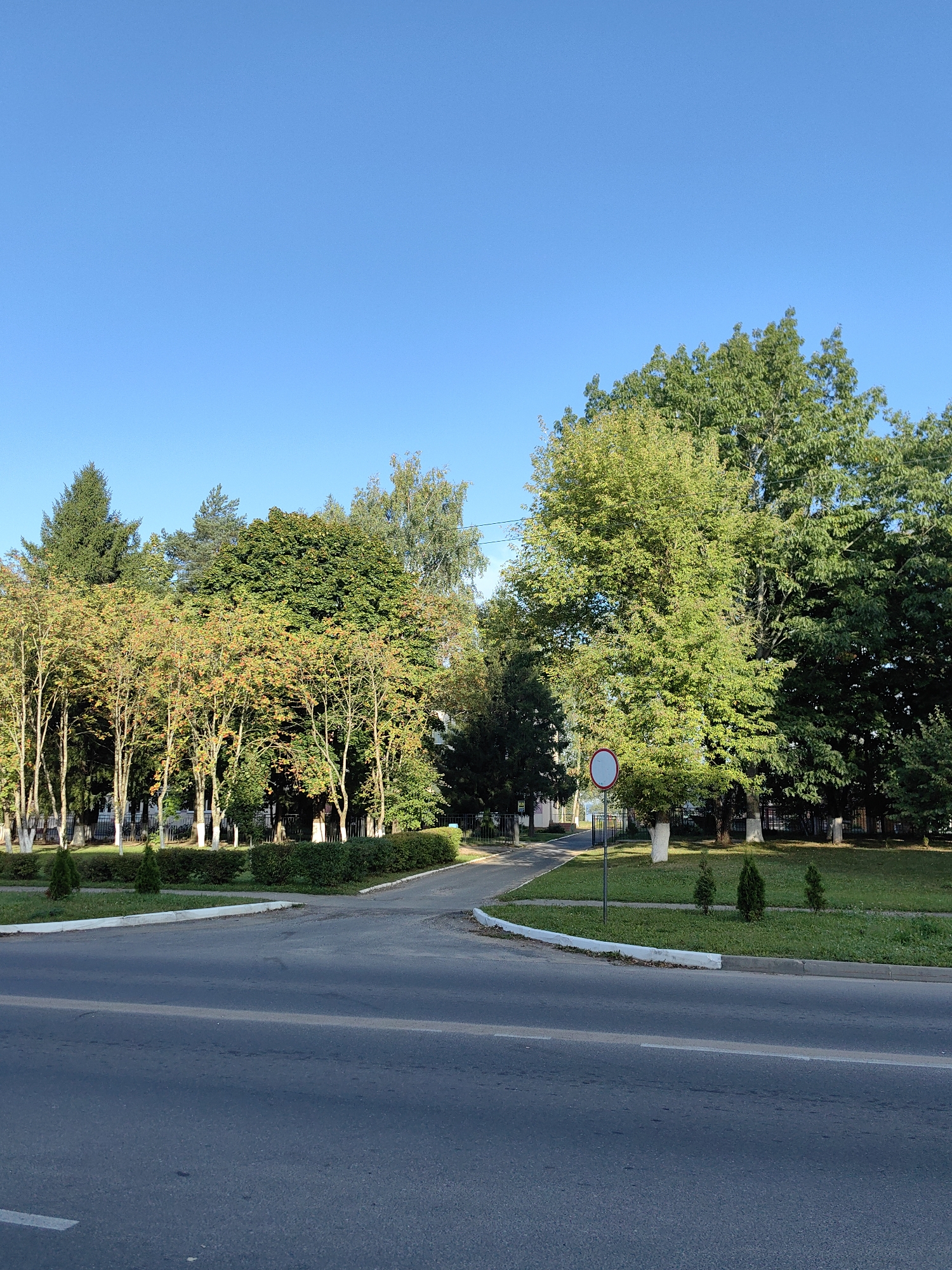 The image depicts an outdoor scene on a clear day, likely in the city of Fanipal, Belarus. In the foreground, a paved road occupies the lower half of the frame, with a white line marking a lane. Beyond the road, a grassy area is bordered by a white curb.
A circular traffic sign with a red border and a white center, indicating a prohibition of some sort, is visible on a pole in the grassy area. A row of neatly trimmed green bushes runs along the edge of the grass.
Behind the bushes, a variety of trees are present. Some trees on the left have sparse foliage with hints of yellow and orange, suggesting it might be autumn. These trees are interspersed with taller, darker green evergreen trees. On the right, a large tree with lush green foliage dominates the background, with more trees extending further into the distance.
The sky above is a clear, bright blue. The overall impression is one of a well-maintained urban green space adjacent to a road.