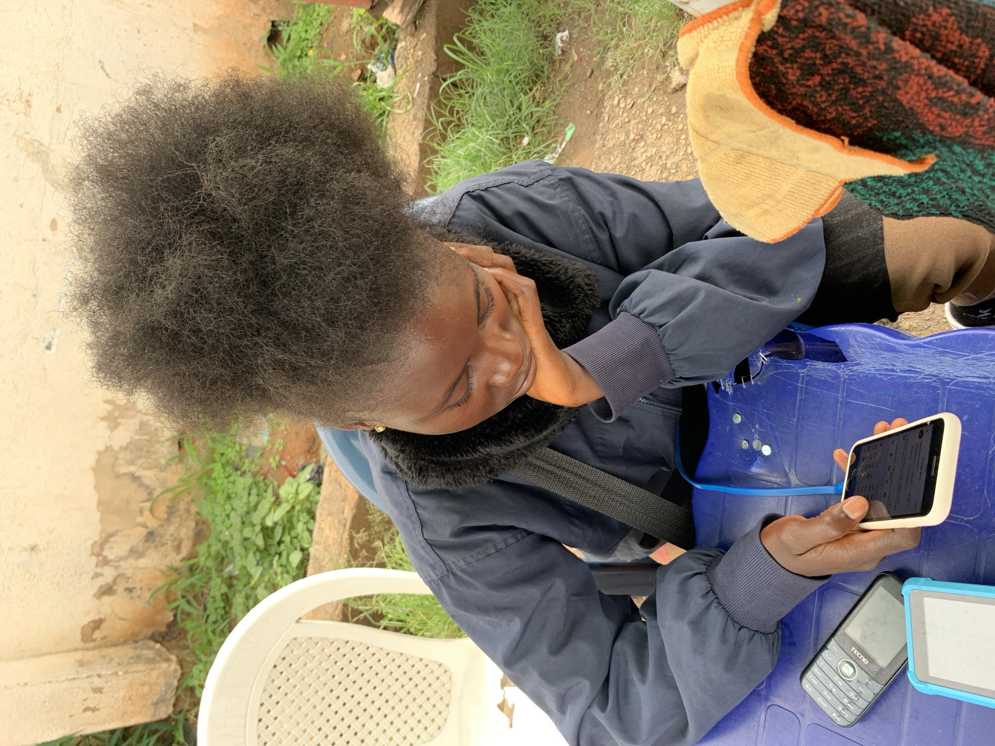 A young African girl with a large, dark afro is seated outdoors, looking down at her smartphone which is connected to a blue charging cable. She rests her chin on her hand, her expression suggesting concentration or contemplation. She wears a dark blue jacket with a fuzzy black collar. A black strap of a bag or accessory crosses her chest. On the bright blue table in front of her are two other electronic devices: a black feature phone and a light blue device with a blank screen. To the right of the table, a colorful textile, possibly a blanket or scarf, is visible. The background shows a weathered, pale-colored wall and green vegetation. The overall setting appears to be an outdoor area, potentially in a more rural or less developed part of Jos, Nigeria, given the visible infrastructure and surrounding environment.