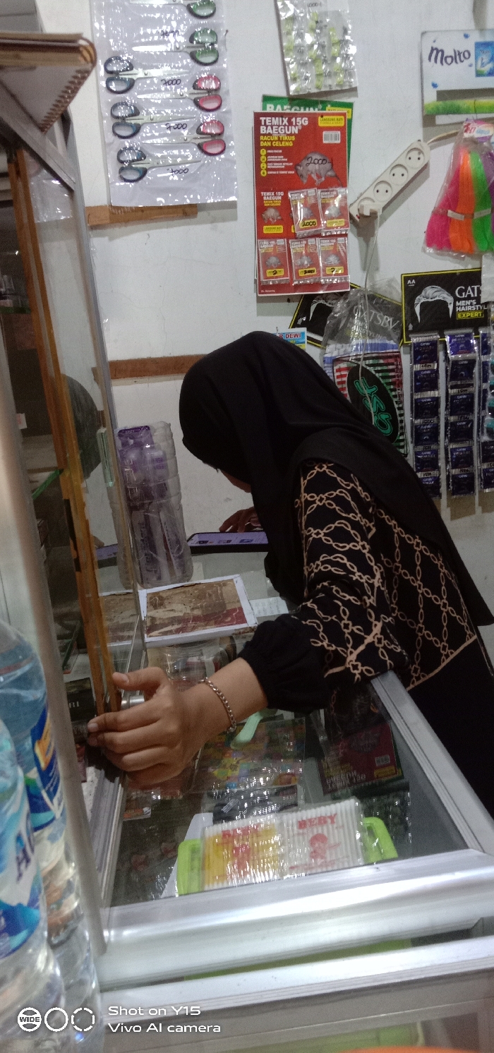 The image depicts an indoor retail setting, likely a small store or kiosk, in Temenggungan, Indonesia. A person, whose upper body is visible from the waist up, is leaning over a glass-topped display case. This individual is wearing a black hijab and a long-sleeved garment featuring a light beige chain-link pattern on a black background. Their right hand, adorned with a thin bracelet, is extended into the open section of the display case, appearing to select or handle an item. A mobile phone displaying a screen is present on the surface of the display case, along with a small stack of papers or cards and a clear plastic bottle containing a purple substance or objects. Inside the display case, a package of "BEBY" brand cotton swabs is clearly visible among other indistinct packaged consumer goods.
The white wall behind the individual serves as a display for various products. Items visible include blister packs of scissors with "7000" written on them, "TEMIX 15G BAEGUN" brand rat poison featuring images of rats and pigs, and smaller packets priced at "2000." A white multi-outlet power strip with five sockets is mounted on the wall. Also displayed are "Molto" brand fabric softener, "GATSBY MEN'S HAIRSTYLING EXPERT" products, and other small packaged items. In the foreground, partially visible on the left, is a plastic bottle of "Aqua" branded water. The overall environment suggests a functional commercial space.
