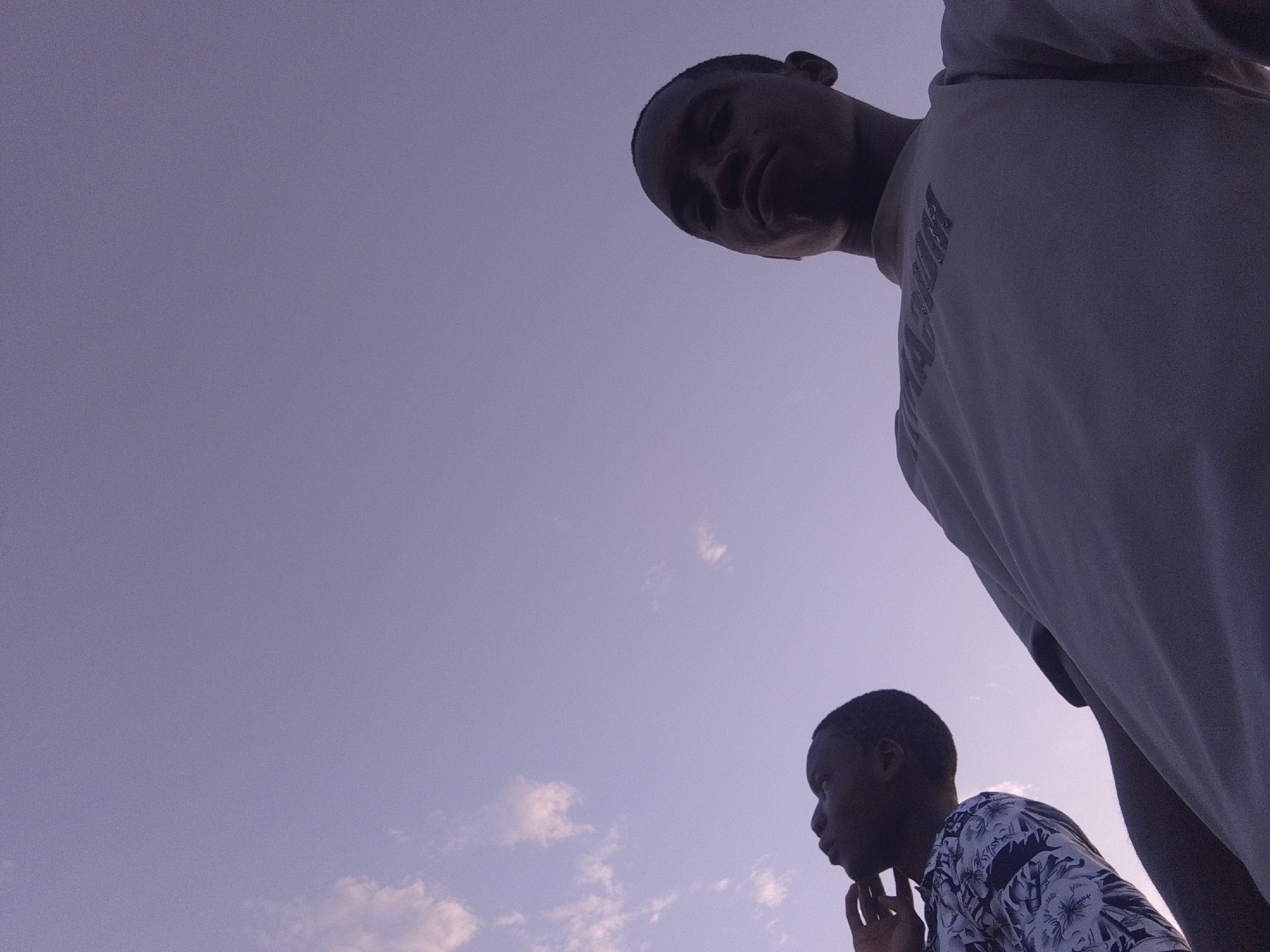 Two young men are present in the image against a light blue sky with scattered clouds. One man is positioned in the lower right of the frame, wearing a shirt with a floral pattern and resting his chin on his hand. The second man is positioned in the upper right, looming over the other, wearing a light-colored t-shirt. No evidence to ascertain the city or country can be obtained from the image.