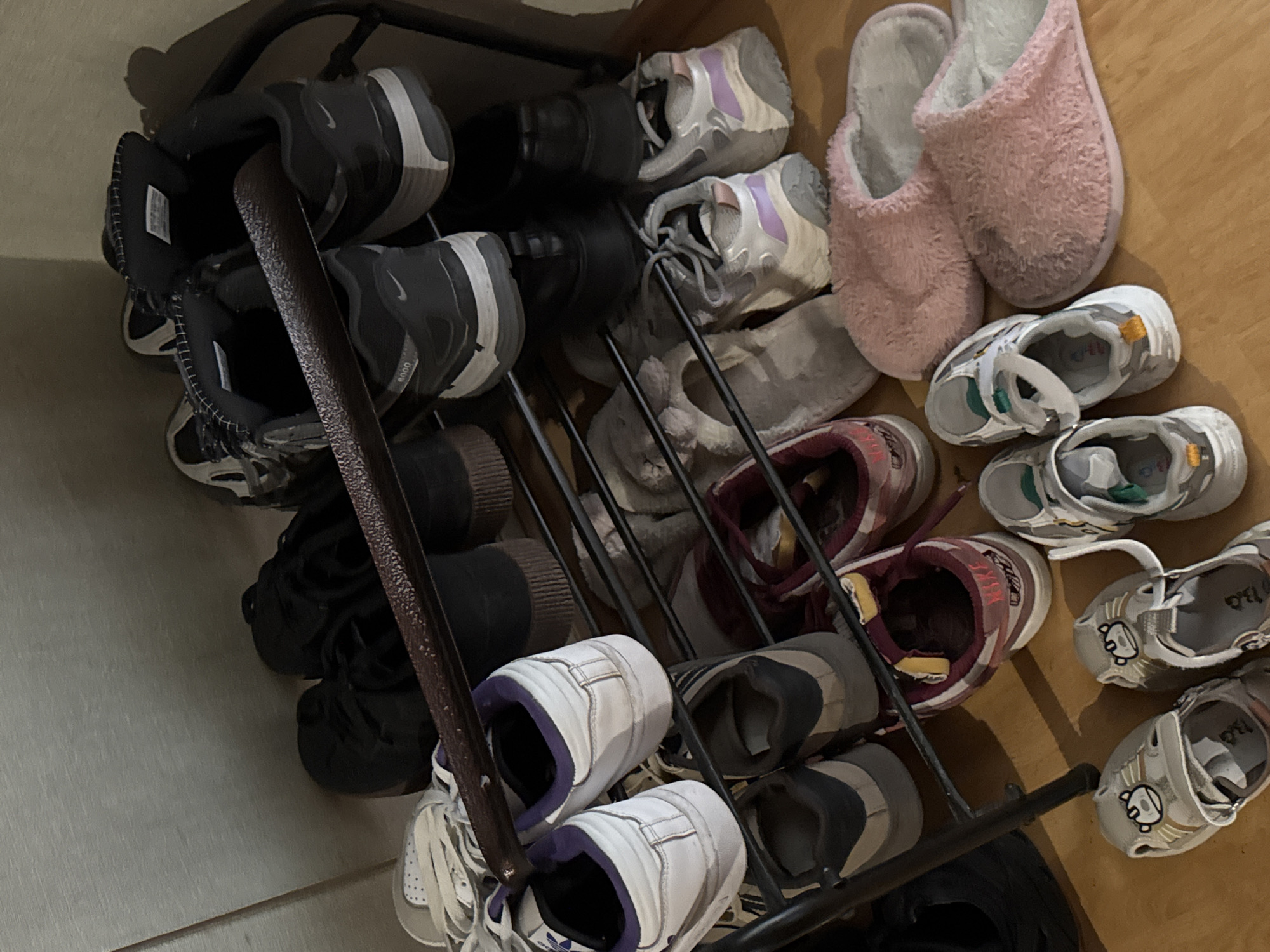 This is a well-lit, medium shot of a multi-tiered shoe rack filled with various types of footwear. The shoe rack is made of dark metal and stands against a plain, light-colored wall. The bottom tier of the rack is on the floor, and the shoes on it are directly on the wooden surface.
The shoes are arranged in pairs, with some placed upright and others on their sides. The collection includes athletic sneakers, casual shoes, and what appear to be slippers. Many of the shoes are dark-colored, such as black and dark grey, but there are also white, grey, and pink options visible. Some of the athletic shoes have visible brand logos, including Nike. The slippers on the bottom are fluffy and appear to be pink and light grey.
The image does not contain any people, and there are no specific actions or interactions to describe. The environment appears to be an indoor space, likely a home, given the shoe rack and the domestic nature of the items. The lighting is even and seems to originate from above and to the right, casting subtle shadows. The overall scene is orderly, with shoes neatly placed on the rack.