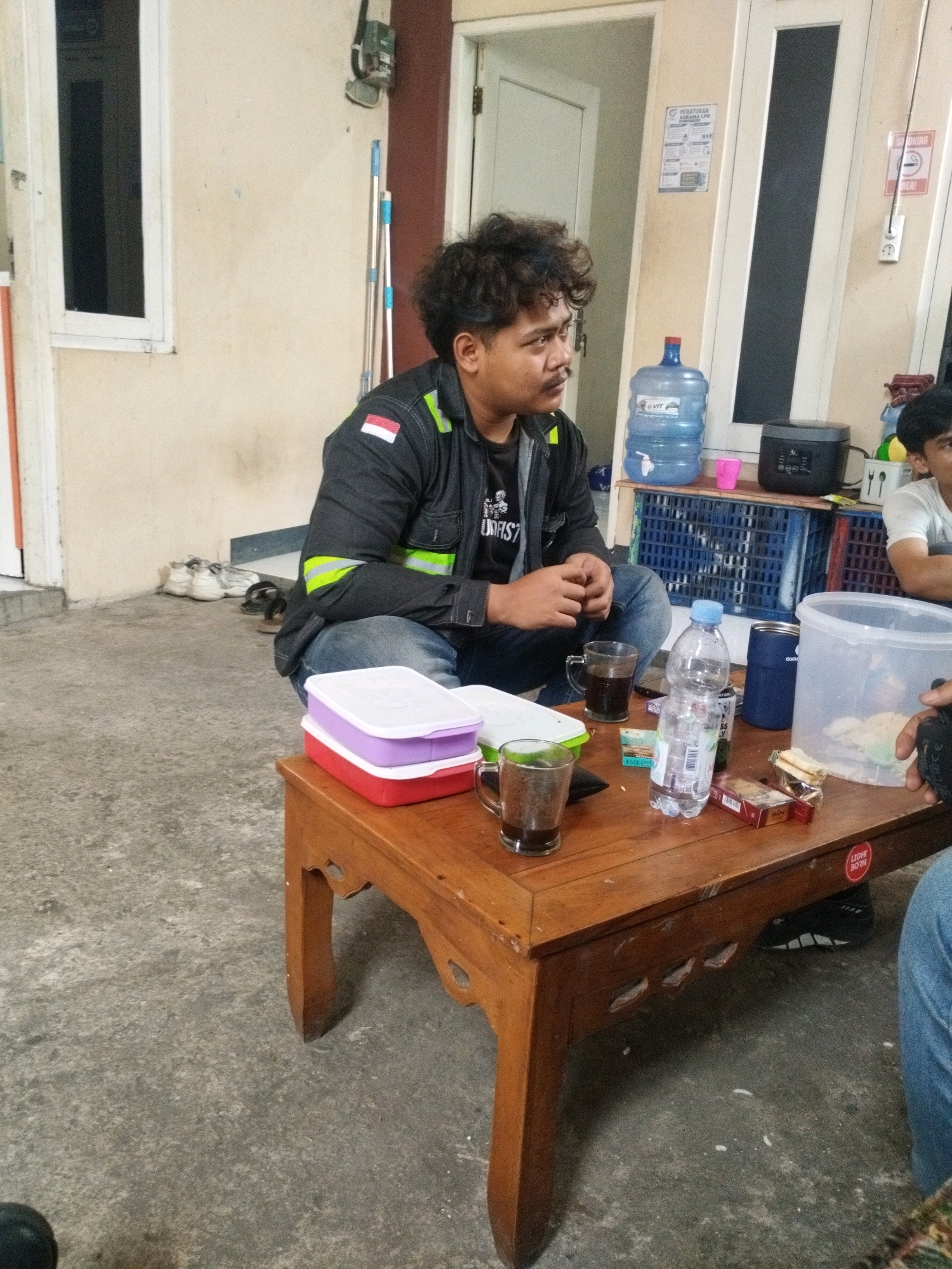 The image depicts an outdoor scene, possibly a communal area or veranda, in Selomerto, Indonesia. A man with curly dark hair is seated cross-legged on the ground, facing a low wooden table. He is wearing a dark jacket with fluorescent green and white stripes on the sleeves and a patch of the Indonesian flag on the left chest. He is looking to his right, with his hands clasped in front of him. On the table are several items, including two glasses with dark liquid, a plastic water bottle, a pack of cigarettes, a sandwich, and a large clear plastic container. Two food containers, one red and one purple, are stacked on the left side of the table. In the background, a water dispenser with a blue bottle is visible, as well as a black rice cooker and some blue plastic crates. A doorway with a white door is on the right, and a window with a white frame is on the left. The ground is made of concrete.
