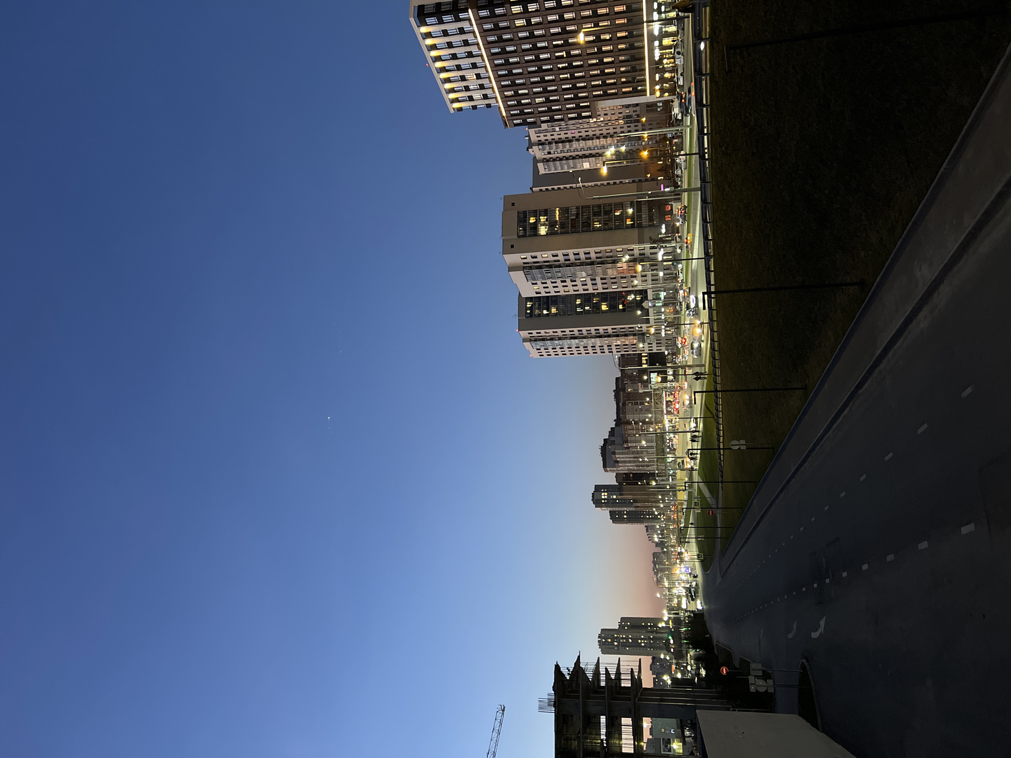 This media file captures a contemporary urban landscape at twilight or early night, likely in Kazan, Russia, given the provided context. The sky, occupying the upper portion of the frame, transitions from a deep, clear blue at its zenith to a lighter blue with subtle hints of orange and pink near the horizon, indicative of sunset or early evening. A faint crescent shape, possibly the moon, is visible in the upper left sky.
Along the left side, a series of modern high-rise apartment or office buildings dominate the cityscape. Many of their windows are illuminated, creating a pattern of warm, glowing rectangles against the darker building facades. These structures extend into the distance, forming a continuous urban fabric. In the foreground on the far left, the incomplete skeleton of a building with visible rebar and scaffolding, accompanied by a tall construction crane, signifies ongoing development.
A wide, dark asphalt road, potentially a multi-lane highway or main street, runs along the right side of the image. It features distinct white dashed lane markings and directional arrows. A narrow strip of dark green grass or landscaping separates the buildings from the road. Numerous streetlights cast a bright, warm light, illuminating the thoroughfare and adjacent areas, contributing to the city's nighttime glow.
No specific activities or individual people are clearly discernible, suggesting a tranquil urban scene after dark, though the illuminated windows and distant vehicle lights imply ongoing city life. No legible text is visible within the image.