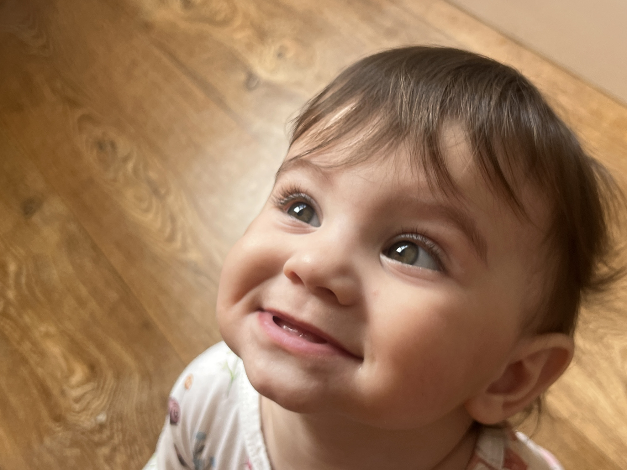 A light-skinned infant or young toddler, with dark brown hair and green eyes, is depicted in a close-up, top-down perspective, looking upwards and to the right with a wide smile, revealing two upper incisor teeth. The child wears a white garment adorned with light-colored floral or leaf patterns. The background consists of a light-brown wooden floor with a visible grain pattern. The scene is located in Elena, Bulgaria.