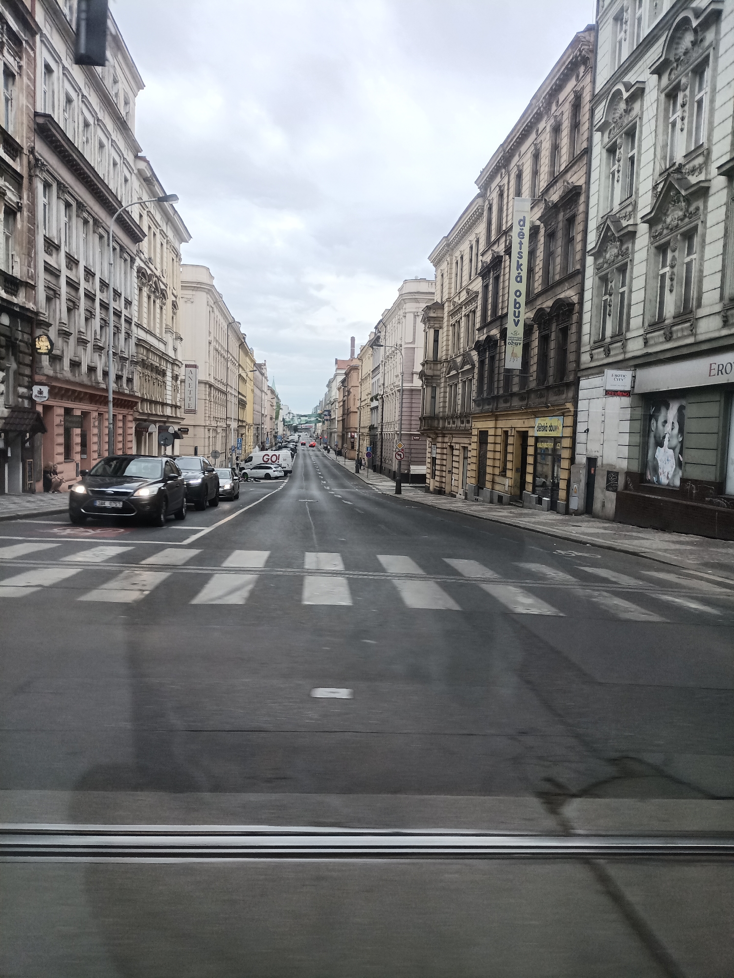 The image shows a street scene in Prague, Czechia, with tall, old buildings lining both sides of the road. The sky is overcast, suggesting a cloudy day. In the foreground, there is a crosswalk with cars stopped at it. The buildings appear to be a mix of residential and commercial spaces, with shops and advertisements visible on the ground floors. One banner reads "Dětská obuv" which translates to "Children's shoes" in Czech. Another sign on the right building says "EROTIC CITY". The street is paved, and there are streetlights and traffic signals visible. The overall impression is of a busy urban environment.