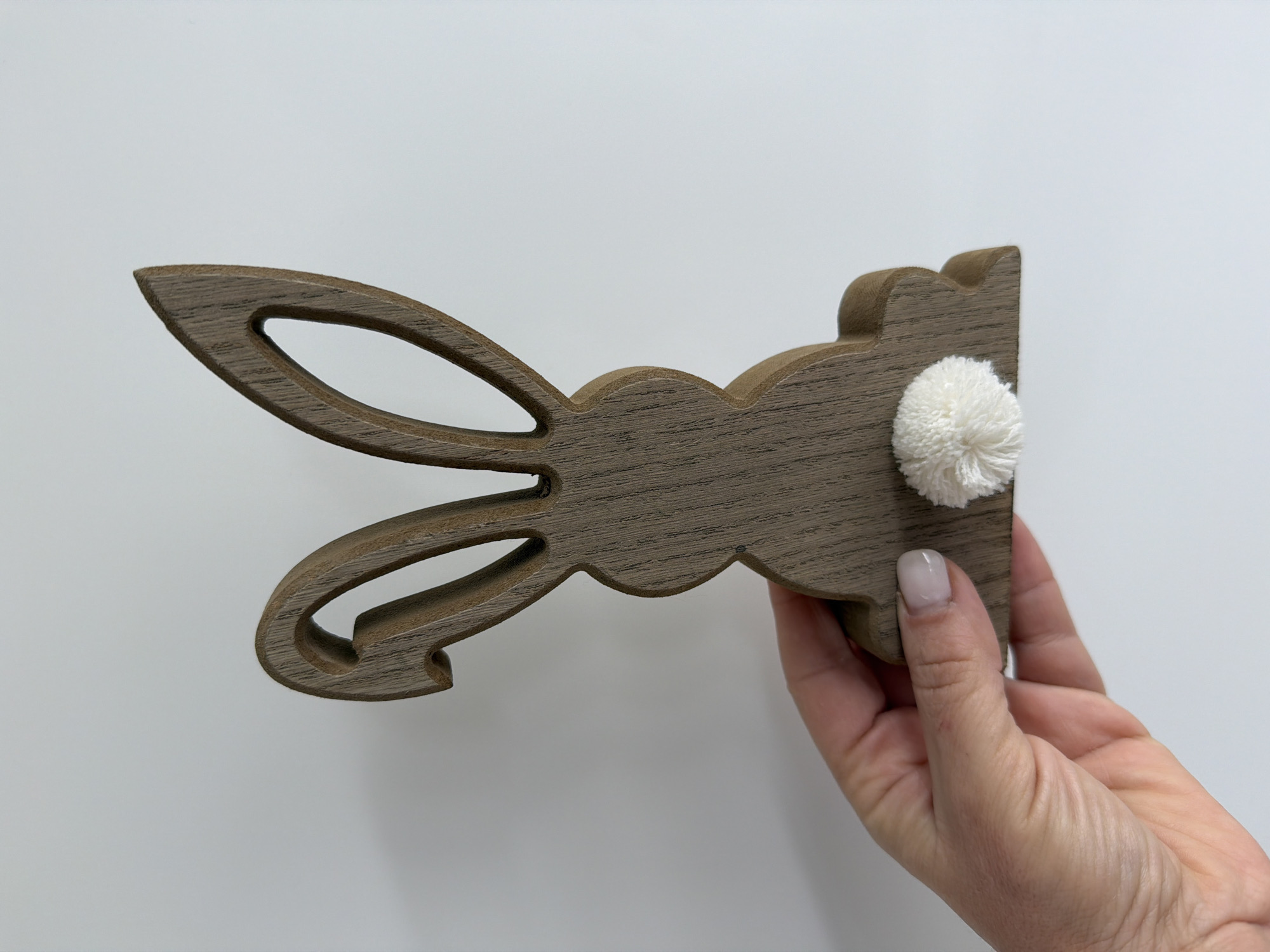 A person's hand holds a brown, wooden bunny decoration against a white background. The decoration consists of bunny ears and the top of the bunny's head, with a fluffy white pom-pom representing the tail. The environment is a plain white surface, providing a neutral backdrop for the subject. Junglinster, Luxembourg is not apparent.