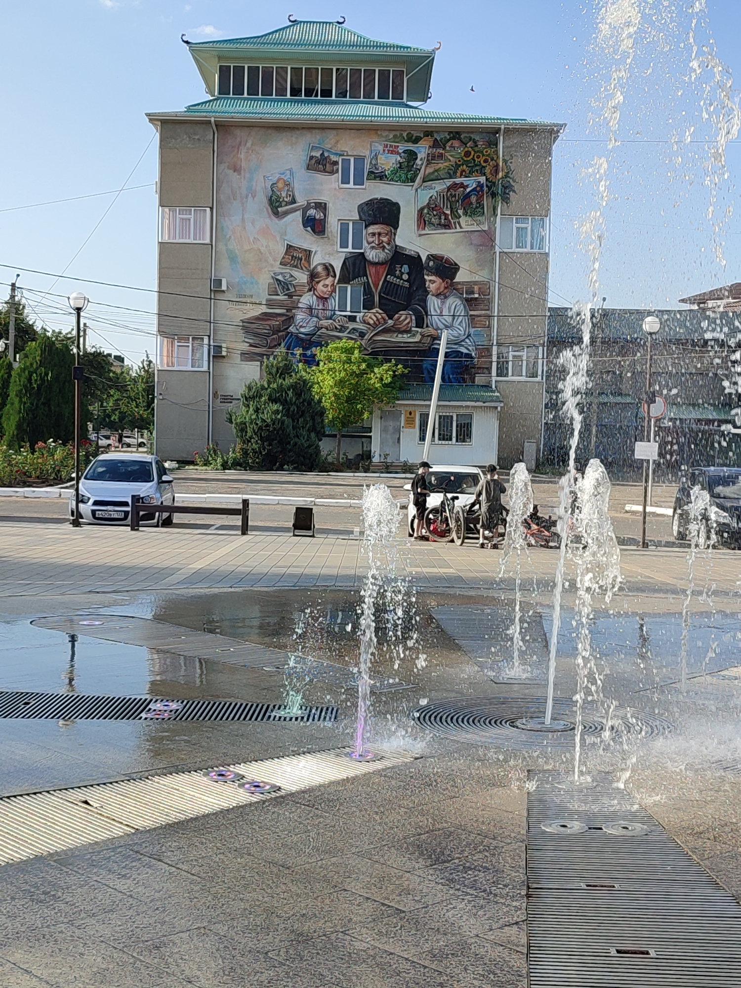 The image depicts a street scene in the city of Abinsk, Russia, featuring a large mural on the side of a building. The mural portrays an elderly man in traditional attire, with medals on his chest, reading a book to a young boy. Above them, several framed images are depicted, including scenes of horses, a soldier, and sunflowers. The building itself has a distinctive roof with Asian architectural elements.
In the foreground, a modern fountain with spraying water is visible, casting reflections on the paved ground. Several vehicles, including a silver car and two bicycles leaning against a white car, are parked on the street. The environment appears to be an urban setting with trees, lampposts, and power lines. The sky is clear and blue.