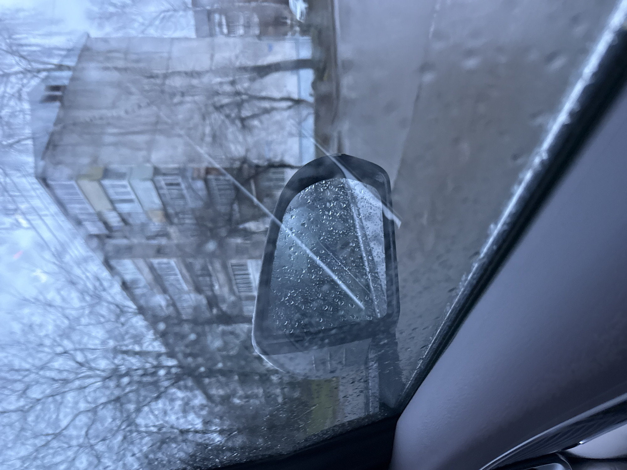 The image shows a rainy day view from inside a car, looking out through the windshield. The car's side mirror is visible on the left. Through the rain-streaked glass, a building is visible, along with bare tree branches. The interior of the car is dark and out of focus. It is not possible to determine the location in Chaikovsky, Russia.
