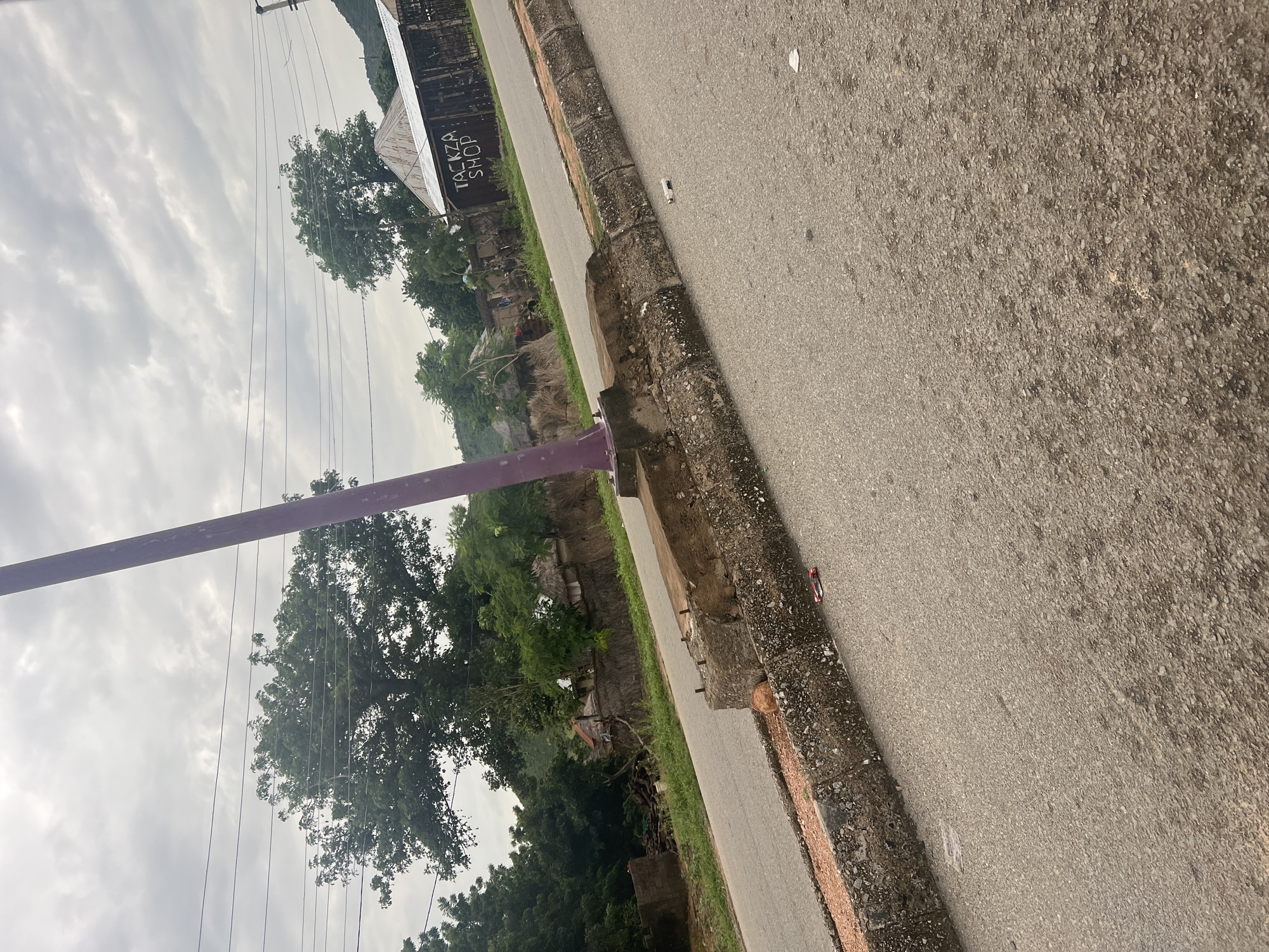 The image depicts an outdoor scene in Kona Garu, Nigeria, with a focus on a roadside and surrounding vegetation under an overcast sky. A paved road runs diagonally across the frame, bordered by a textured concrete curb. To the left of the road, lush green trees and foliage create a dense backdrop, with a few structures partially visible amidst the greenery. One building has a weathered, corrugated metal roof with visible text reading "TACKZA SHOP." A purple pole stands prominently in the foreground, extending vertically. Power lines crisscross the sky, suggesting an inhabited area. The overall mood is muted, likely due to the cloudy weather, which indicates it could be daytime but not particularly sunny. There are no people clearly visible in the frame, and the scene appears to be one of everyday life in a semi-rural or suburban setting.