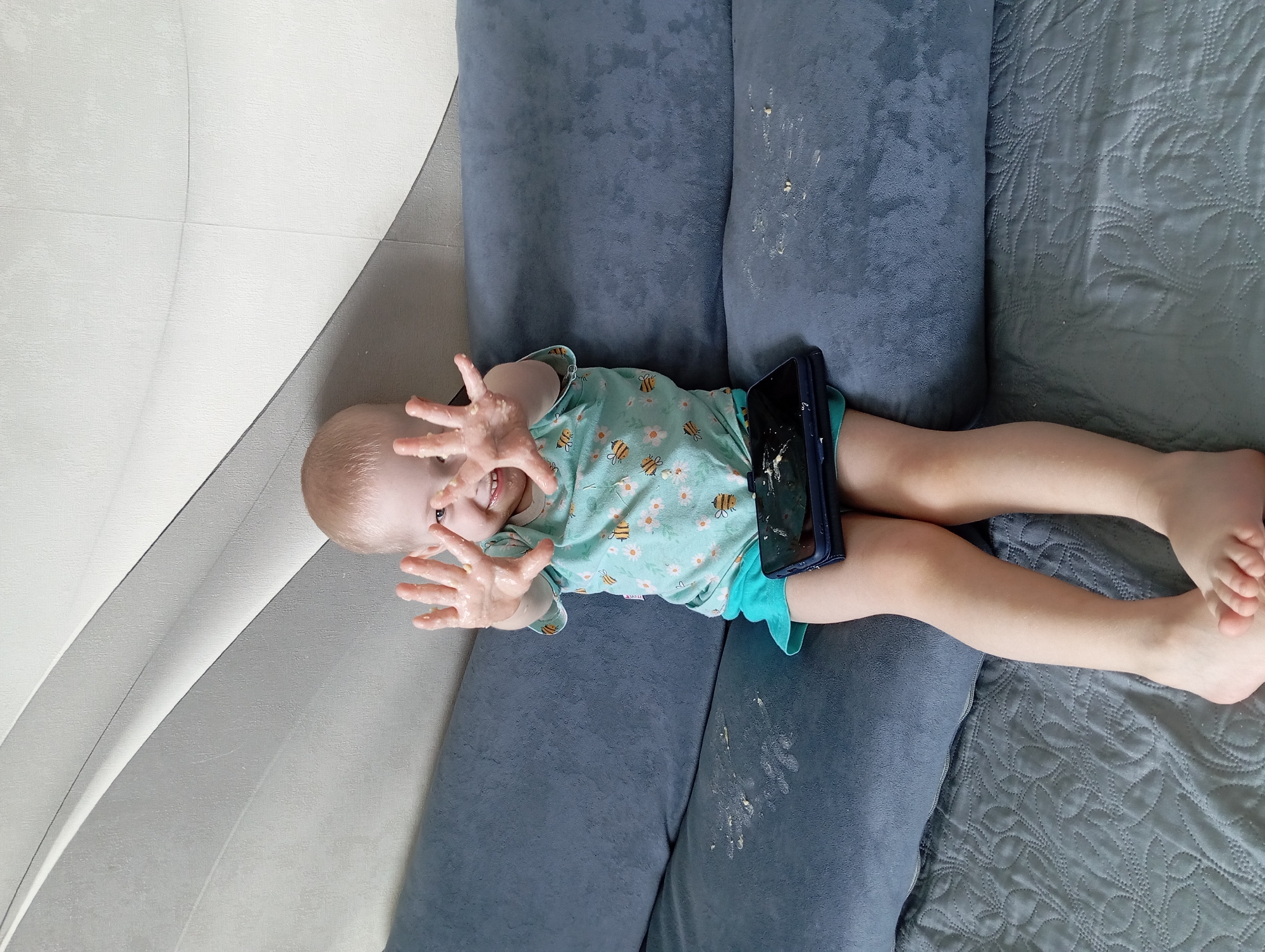 A young child is captured in an overhead shot, lying horizontally on a blue fabric sofa. The child, likely a toddler, has light hair and is wearing a light blue/teal t-shirt with bumblebee and daisy patterns, and matching teal shorts. They are looking directly at the camera with a wide smile, and both hands are raised, palms open, appearing covered in a light-colored, sticky substance, possibly pureed food.
A dark-colored smartphone, possibly in a case, rests on the child's stomach area, also showing some of the sticky substance on its screen and case. The blue sofa cushion beneath the child's torso also has visible smudges and streaks of the same food-like substance. To the right of the child, a grey quilted blanket with an intricate pattern covers part of the surface. To the left, a light-colored wall with a subtle textured pattern and a curved design is visible. The lighting is bright and even, suggesting it is daytime indoors. The scene conveys a playful moment, possibly after a messy meal or snack, set in a home in Yurgamysh, Russia. No visible text is present.