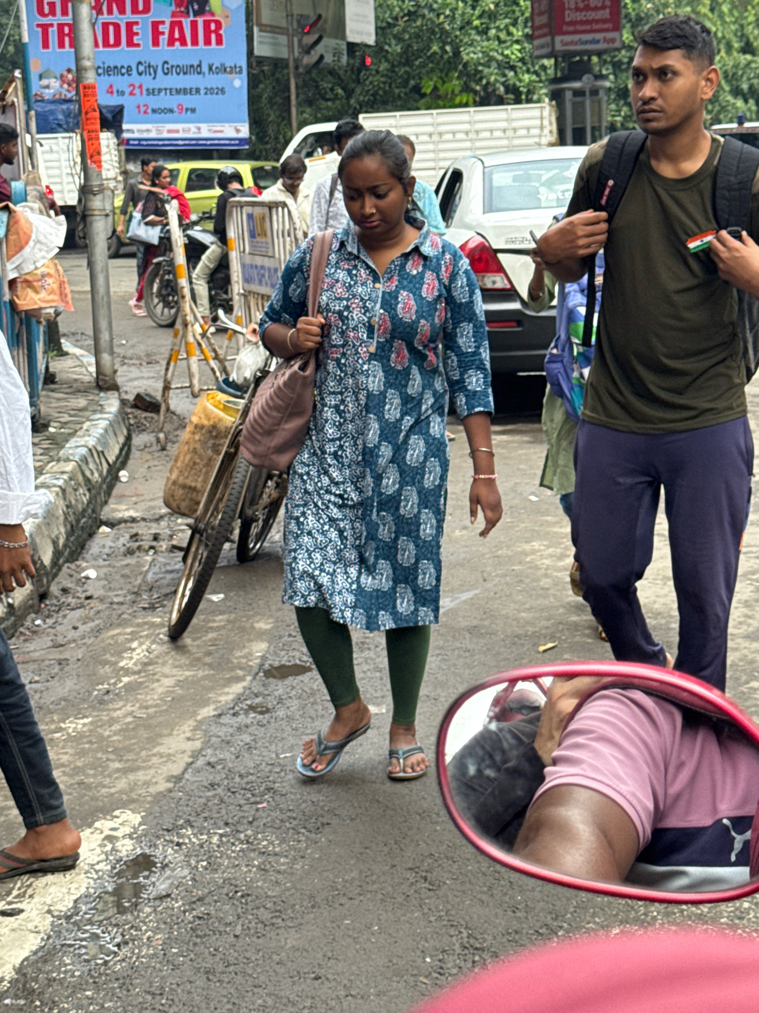 In Kolkata, India, a street scene unfolds with pedestrians and vehicles. A woman in a blue patterned dress and green leggings walks forward, carrying a tan bag. To her right, a man in a dark green t-shirt and navy pants walks, wearing a backpack. In the background, a yellow vehicle and other people are visible. The ground is wet and appears to be a road with some debris. A red side-view mirror in the foreground reflects a person wearing a pink shirt. A large blue banner in the background advertises a "GRAND TRADE FAIR" at "Science City Ground, Kolkata" from September 4th to 21st, 2026.