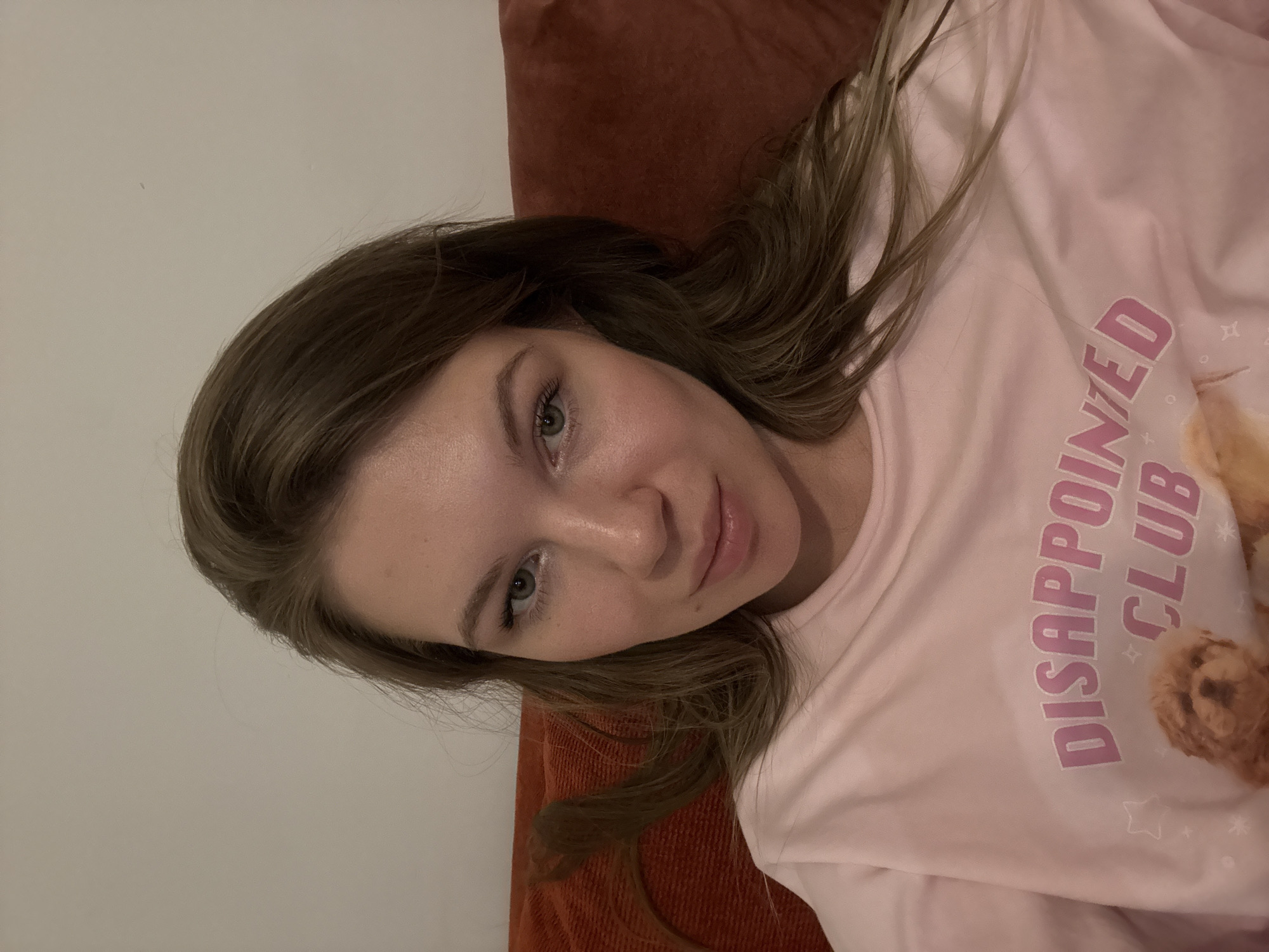 A young woman reclines indoors, looking directly at the camera with a soft, gentle smile. She has long, wavy light brown hair that cascades around her shoulders and over a reddish-brown textured surface, likely a pillow or sofa arm. Her eyes are light-colored, and she appears to have minimal makeup, with visible mascara.
She is wearing a light pink long-sleeved top. Prominently displayed on the front of her shirt are the words "DISAPPOINTED CLUB" in bold pink lettering, accompanied by an illustration of a fluffy, light brown dog. Small star and plus symbols are also scattered across the fabric of the shirt. The background consists of a plain, light-colored wall, indicating an indoor setting, possibly a home in Vozdovac Urban Municipality, Serbia. The lighting suggests a well-lit environment, but no specific time of day can be determined. The overall impression is a casual, relaxed portrait.