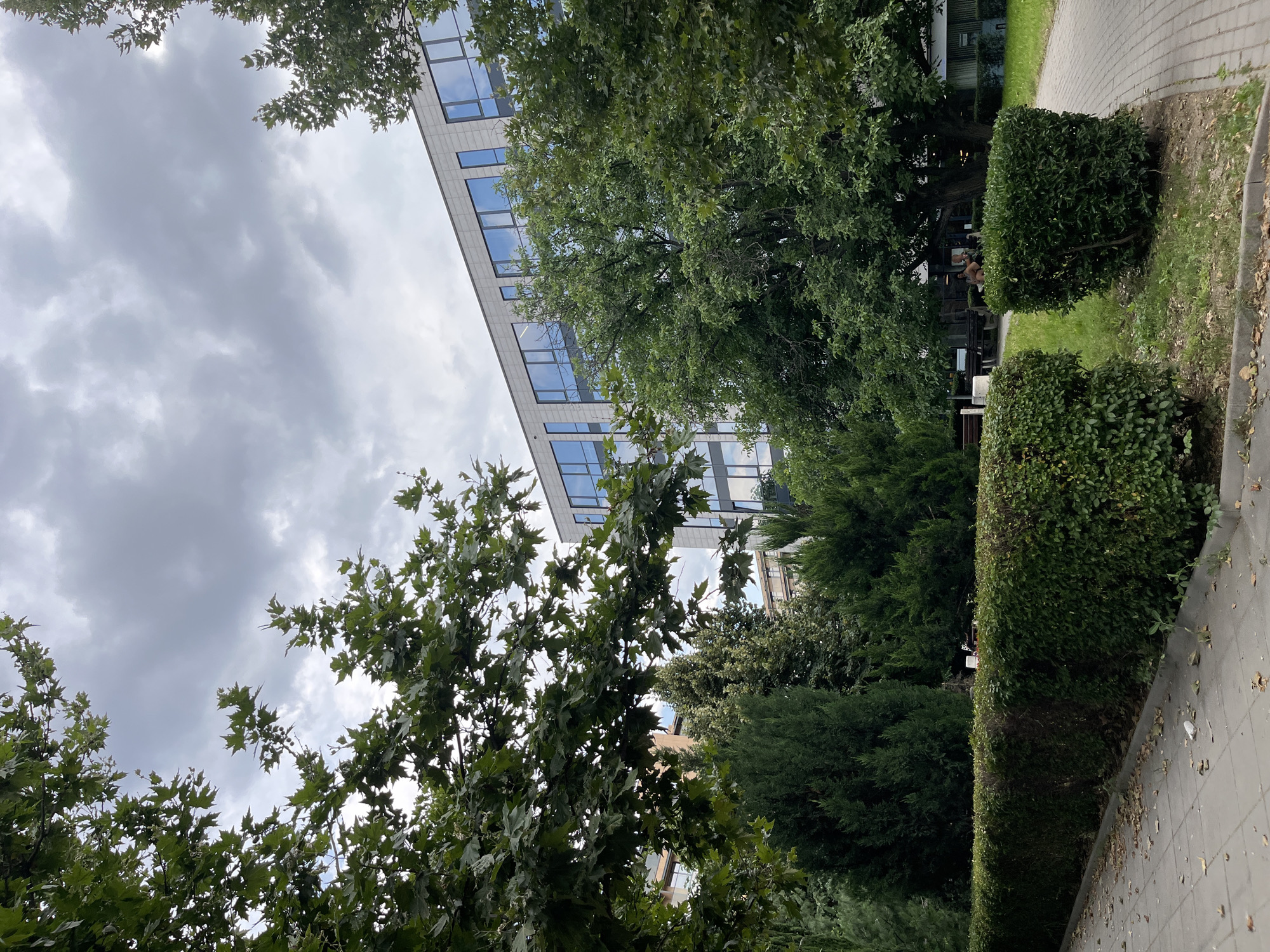 This is an outdoor, eye-level, full shot of a modern building with large blue-tinted windows, partially obscured by lush green trees and manicured bushes. A paved walkway runs along the right side of the frame, leading towards the building. The sky is overcast with heavy, gray clouds. The building appears to be an office or commercial structure, judging by its architecture and the reflective windows. In the foreground, a neat hedge lines the walkway, and fallen leaves are scattered on the pavement. Further into the scene, more trees and bushes create a park-like atmosphere. There are several benches visible in the background, and two individuals are seated on one of them, facing away from the camera. The overall impression is a well-maintained urban green space adjacent to a contemporary building. The architectural style and the vegetation are consistent with urban areas in Plovdiv, Bulgaria.