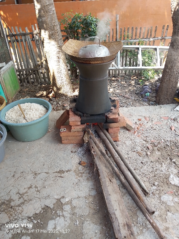 The image shows a traditional cooking method in Gegesik, Indonesia, at coordinates -6.538 latitude and 108.43 longitude. A large, black pot with a metal lid is perched on a brick platform. The pot is nestled within a woven bamboo basket, and steam rises from the pot's opening. The pot sits atop a small fire fueled by wooden logs and a small pile of charcoal. A green plastic basin filled with white rice is visible to the left of the pot. The scene is set in an outdoor area with a wooden fence and a large tree trunk on the left. The ground is a cracked concrete surface. The scene is shot from a low angle, likely taken by a person standing nearby. The photo is stamped with a timestamp: 17 Mei 2025, 10:27.