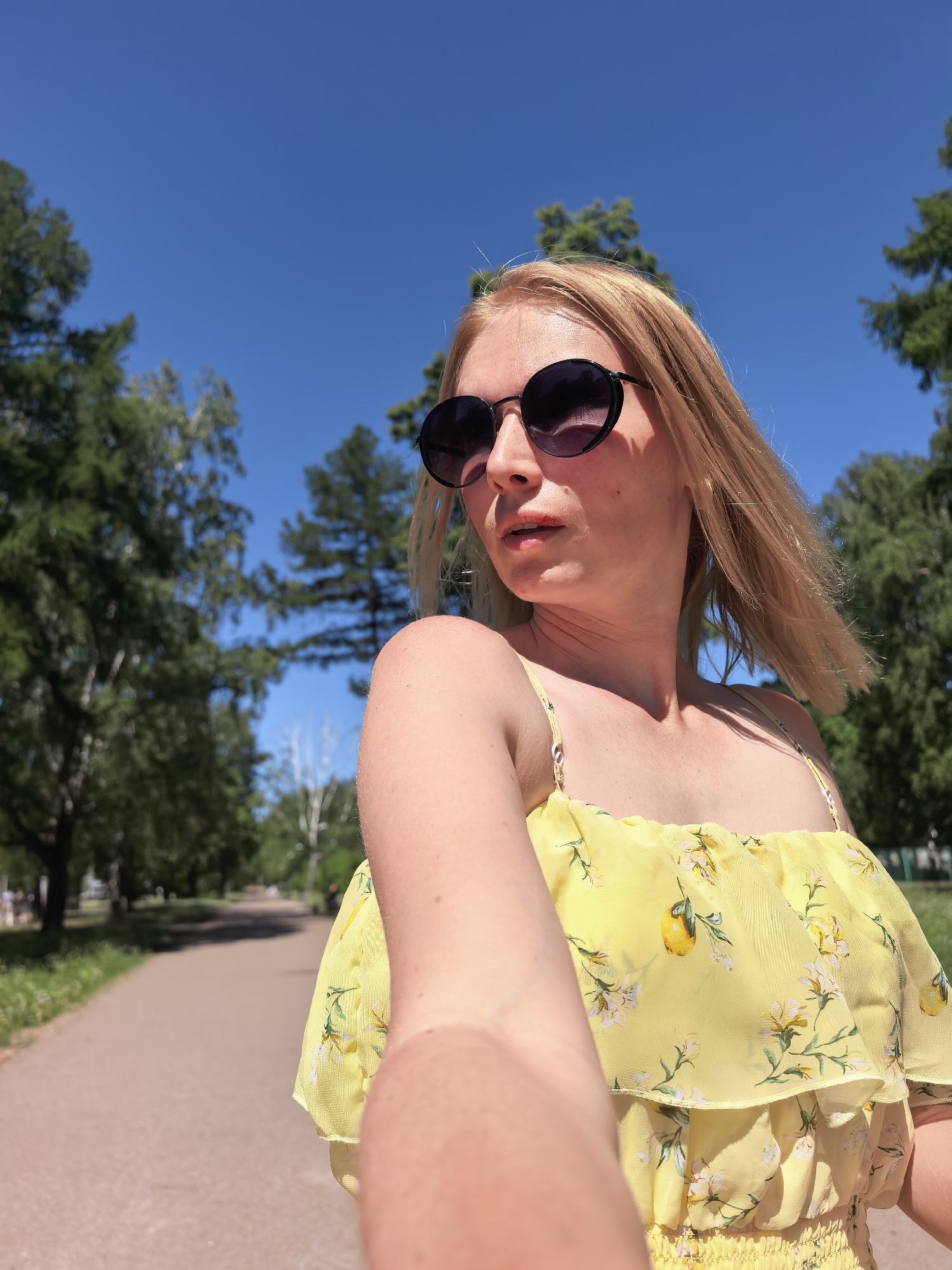 A person wearing sunglasses and a yellow floral dress is taking a selfie on a path in a park. Tall trees line the path on either side. The sky is blue. The person's hair is blonde and blowing in the wind. Based on visual evidence, the location is in a city park, possibly in Sterlitamak, Russia, given the request.