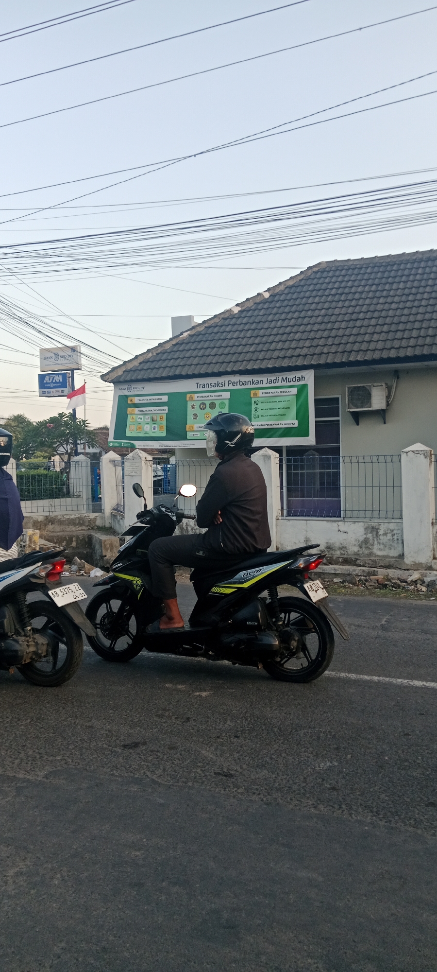 In Maguwoharjo, Indonesia, a person wearing a black helmet and jacket is seated on a black and yellow scooter. The scooter has "BEAT" written on its side. The individual is wearing dark pants and light-colored sandals, with their arms crossed. The scooter is parked on a paved road, and another white scooter is visible to the left. Behind the scooters, there is a building with a tiled roof and a sign that reads "Transaksi Perbankan Jadi Mudah" (Banking Transactions Made Easy). The building also has a white fence and an air conditioning unit mounted on the wall. Overhead, numerous power lines crisscross the sky. A red and white flag, likely the Indonesian flag, is also visible on a flagpole to the left of the building.