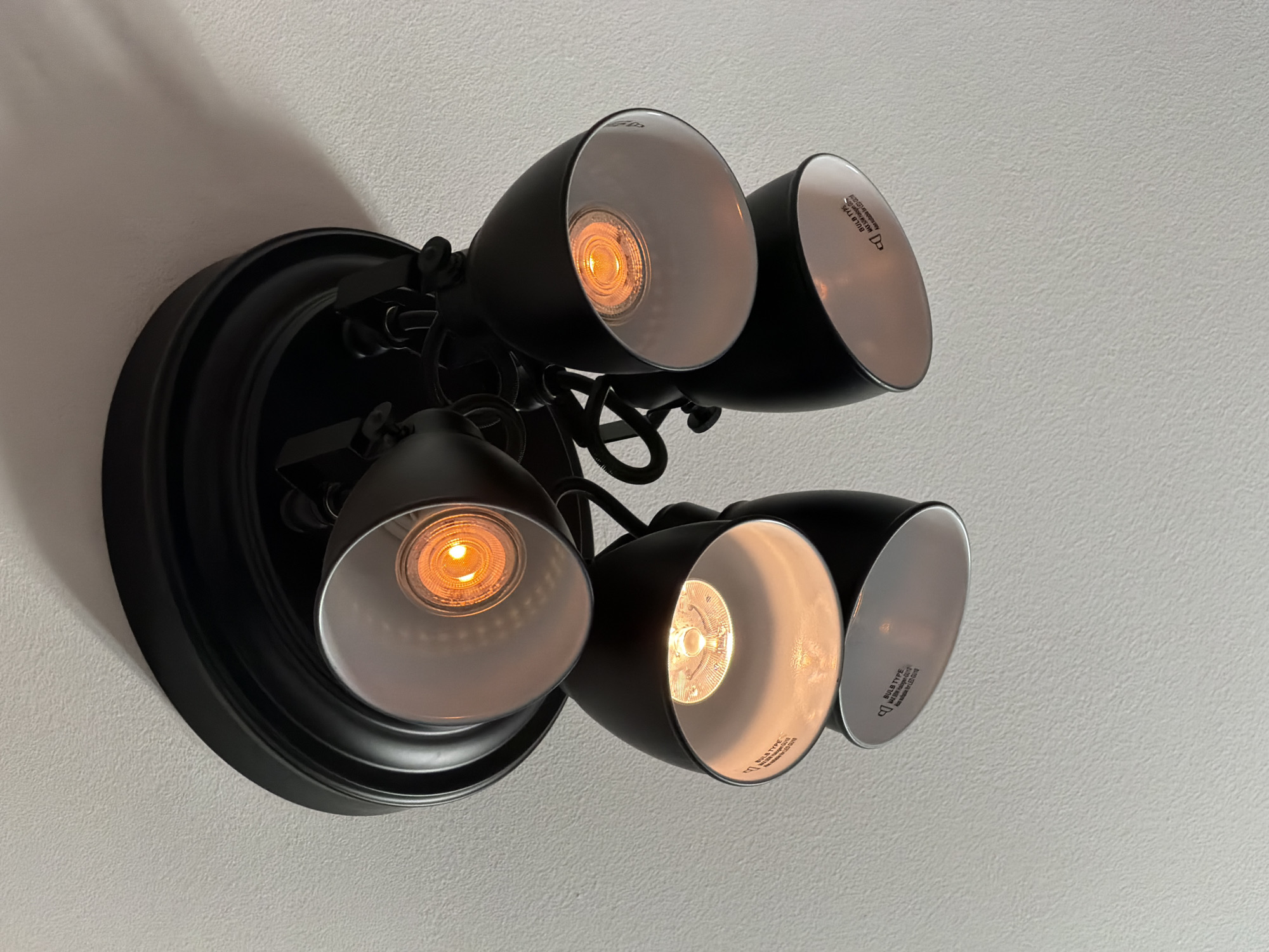 A black ceiling mounted track light fixture with four adjustable spotlights is shown from a low angle against a white textured ceiling. The fixture has a circular base and the spotlights are cone-shaped with white interiors. All four bulbs are illuminated with a warm yellow light. Some of the spotlights have visible text on their inner surfaces, indicating "BULB TYPE" and "MAX 50W Halogen GU10". The image does not depict any people, animals, or identifiable landmarks. The scene is indoors and appears to be a residential or commercial space. There is no information in the image to suggest a specific location in Malmö, Sweden.