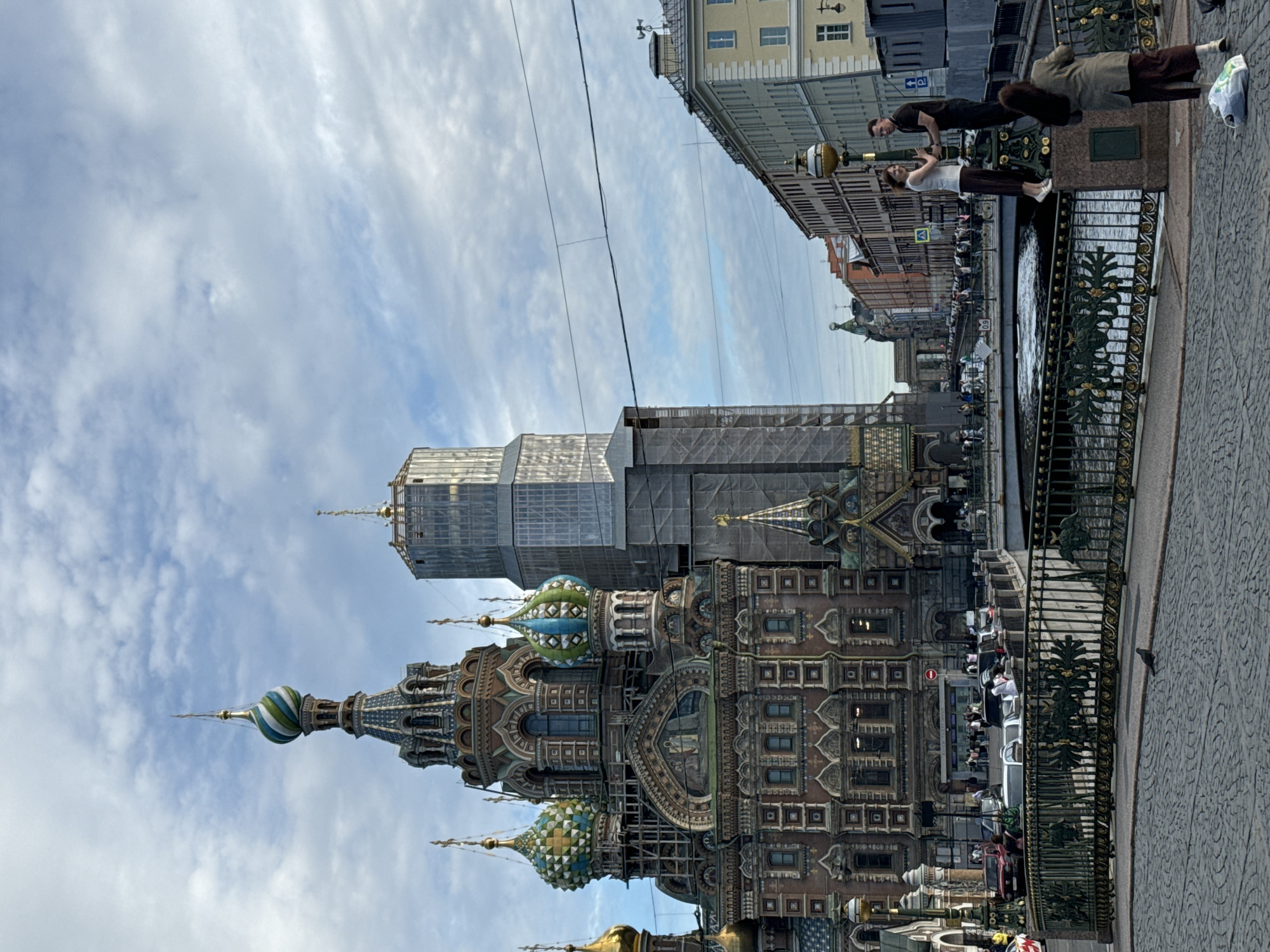 The image shows the Church of the Savior on Spilled Blood in Saint Petersburg, Russia. The church's colorful onion domes and intricate facade are visible, with some sections under scaffolding, suggesting ongoing restoration or construction. In the foreground, a group of people are standing on a bridge overlooking a canal. Two individuals are in the process of a handstand or yoga pose, while others observe or walk by. The scene appears to be set during daylight, with a cloudy sky. The architecture of the surrounding buildings and the ornate metalwork of the bridge railings provide further context for the urban setting of Saint Petersburg.