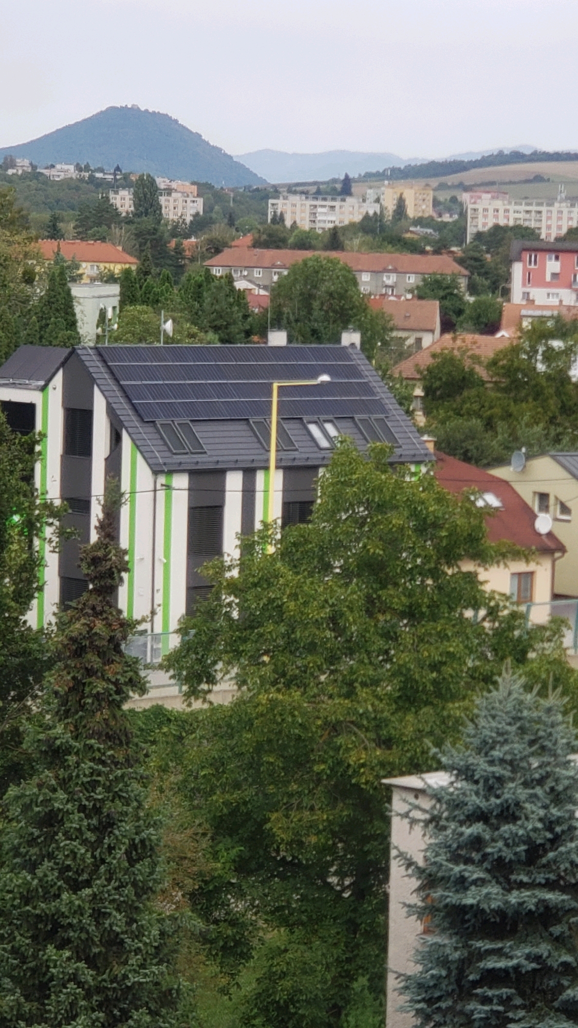 The image displays a residential building with a modern design, characterized by vertical stripes of white and lime green on its facade. The building features a dark-colored roof covered with solar panels and several skylights. A bright yellow streetlamp stands in front of the building. Surrounding the structure is lush greenery, including various trees and evergreens. In the background, a hilly landscape is visible with a mountain in the distance, and beyond that, more rolling hills under a hazy sky. Several other buildings, including apartment blocks and houses with red-tiled roofs, are scattered throughout the urban and semi-urban environment. This scene is located in the city of Prešov, Slovakia, specifically in the Sekčov housing estate, known for its mix of modern and older residential architecture, nestled against a backdrop of natural terrain. There are no people or animals visible in the image.