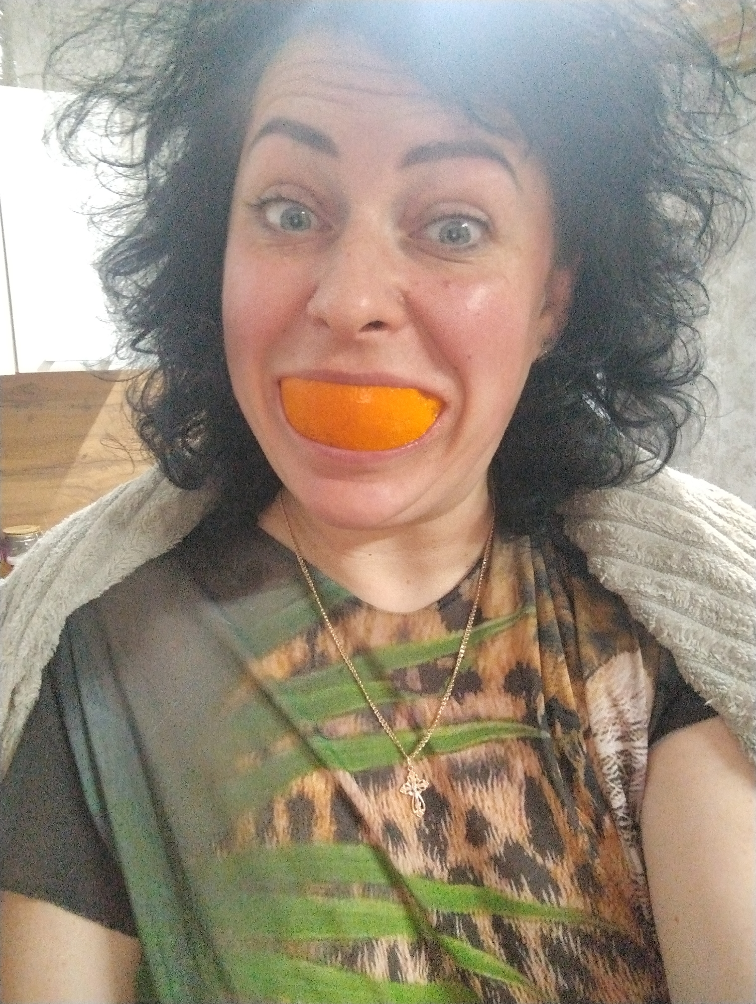 This close-up portrait features a woman with voluminous dark, curly hair and wide, light-colored eyes, conveying a playful or surprised expression. She is holding a slice of orange fruit in her mouth, creating an exaggerated, wide "orange smile."
The woman is wearing a top with a distinctive pattern that combines leopard print with green, leafy motifs. Around her neck, she has a gold chain necklace with a small, detailed cross pendant. A light gray, possibly plush or textured, blanket or shawl is draped over her shoulders.
The setting appears to be indoors, likely a casual home environment, with a softly blurred background that includes light-colored walls or cabinets and hints of wooden surfaces. The lighting suggests it is daytime. The activity is posing for a humorous photograph. No visible text is present in the image. This scene takes place in Grishanki, Russia.