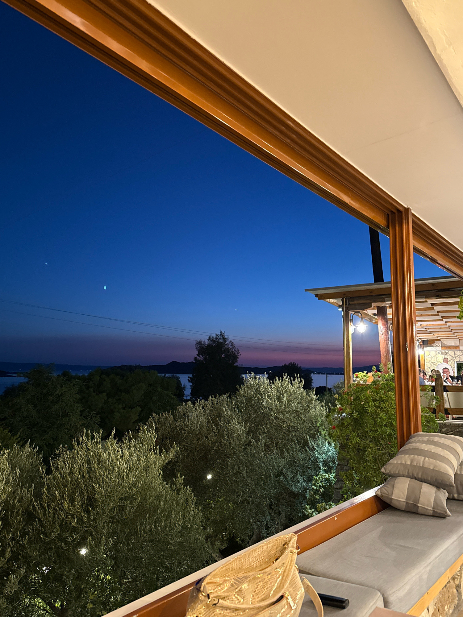 The image depicts a view from an outdoor seating area, likely a balcony or patio, overlooking a body of water and a distant shoreline at dusk. The sky is a deep blue, transitioning to hues of purple and pink near the horizon. Several trees with dense foliage are visible in the foreground and midground. In the distance, islands or peninsulas can be seen against the twilight sky.
To the right of the frame, part of a covered outdoor restaurant or seating area is visible. Wooden beams and a lattice ceiling are apparent, along with hanging light fixtures. Several people are seated at tables, engaged in conversation or dining. One person, likely a server, is wearing a white shirt and appears to be interacting with diners.
The seating area in the foreground features a long, grey upholstered bench with several striped cushions. A woven straw bag rests on the bench, and a dark object, possibly a remote control, is also present. The railing of the seating area is made of wood, and stone construction is visible at the base of the seating area. The scene is set in Ouranoupoli, Greece, as indicated by the context.