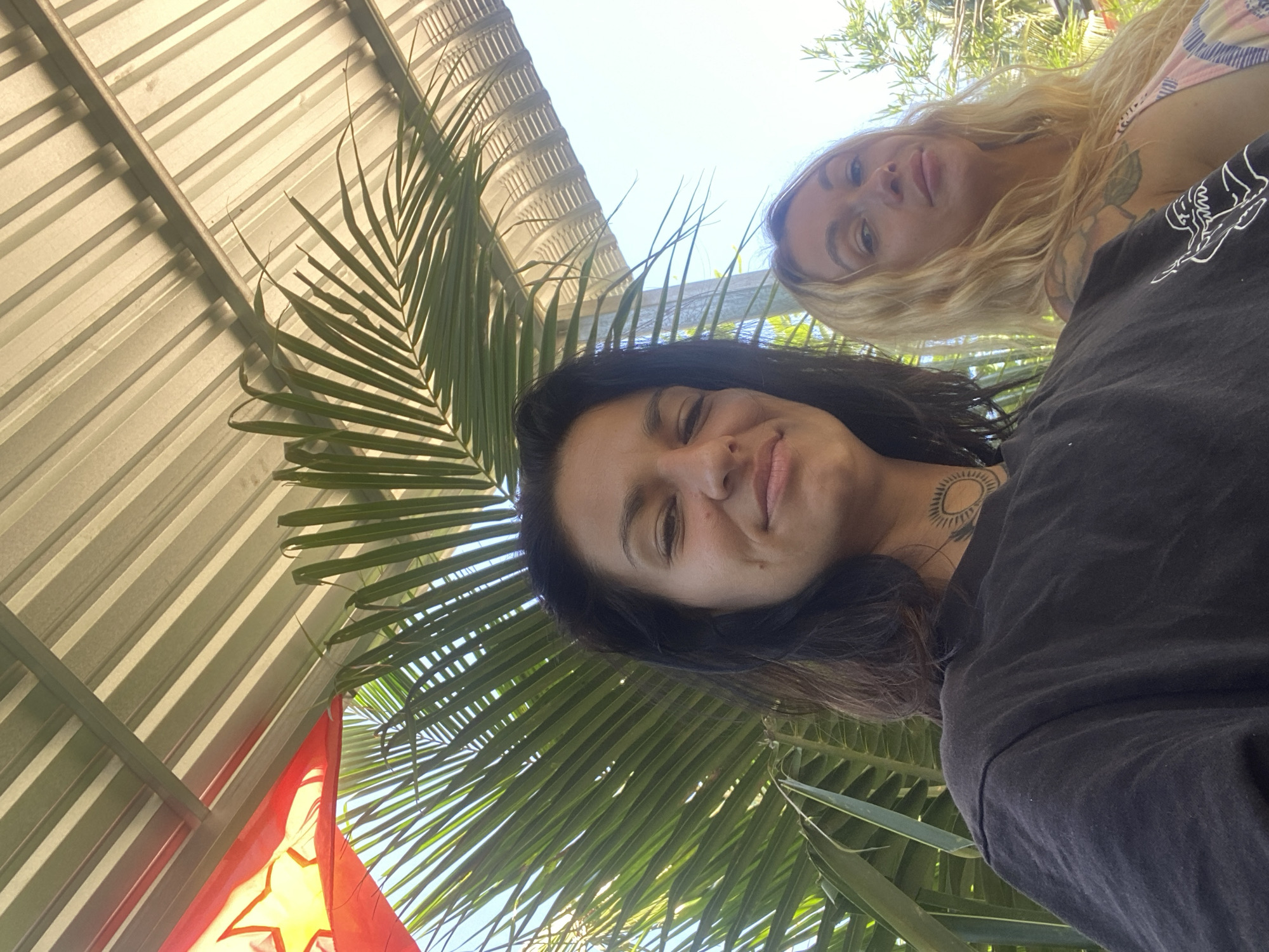 This is a low-angle, outdoor, selfie-style photograph taken in Mũi Né, Vietnam, under a corrugated metal roof structure, possibly a shelter or veranda. A large palm frond dominates the foreground, partially obscuring the two women and the sky. The woman on the left has dark hair and a tattoo of a sunburst on her neck. She is wearing a black t-shirt. The woman on the right has blonde hair and is wearing a patterned top. Both are looking towards the camera. In the upper left corner, a portion of a red flag with a yellow star is visible, possibly belonging to the Vietnamese flag or a decorative item. The overall lighting suggests it is daytime, with sunlight filtering through the palm fronds and casting shadows on the roof. The background shows glimpses of foliage and the sky.