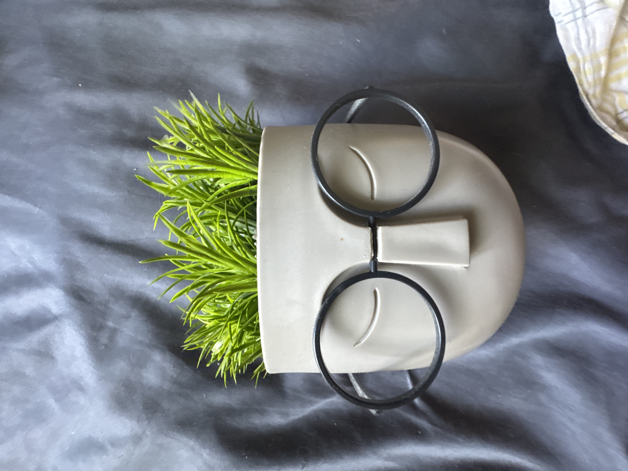 A light gray ceramic planter, shaped like a face with closed eyes and a nose, sits on a dark gray textured surface, possibly fabric. The planter has black wire spectacles perched on its "nose." Inside the planter, bright green artificial grass sprouts upwards. The image is taken from a top-down perspective.