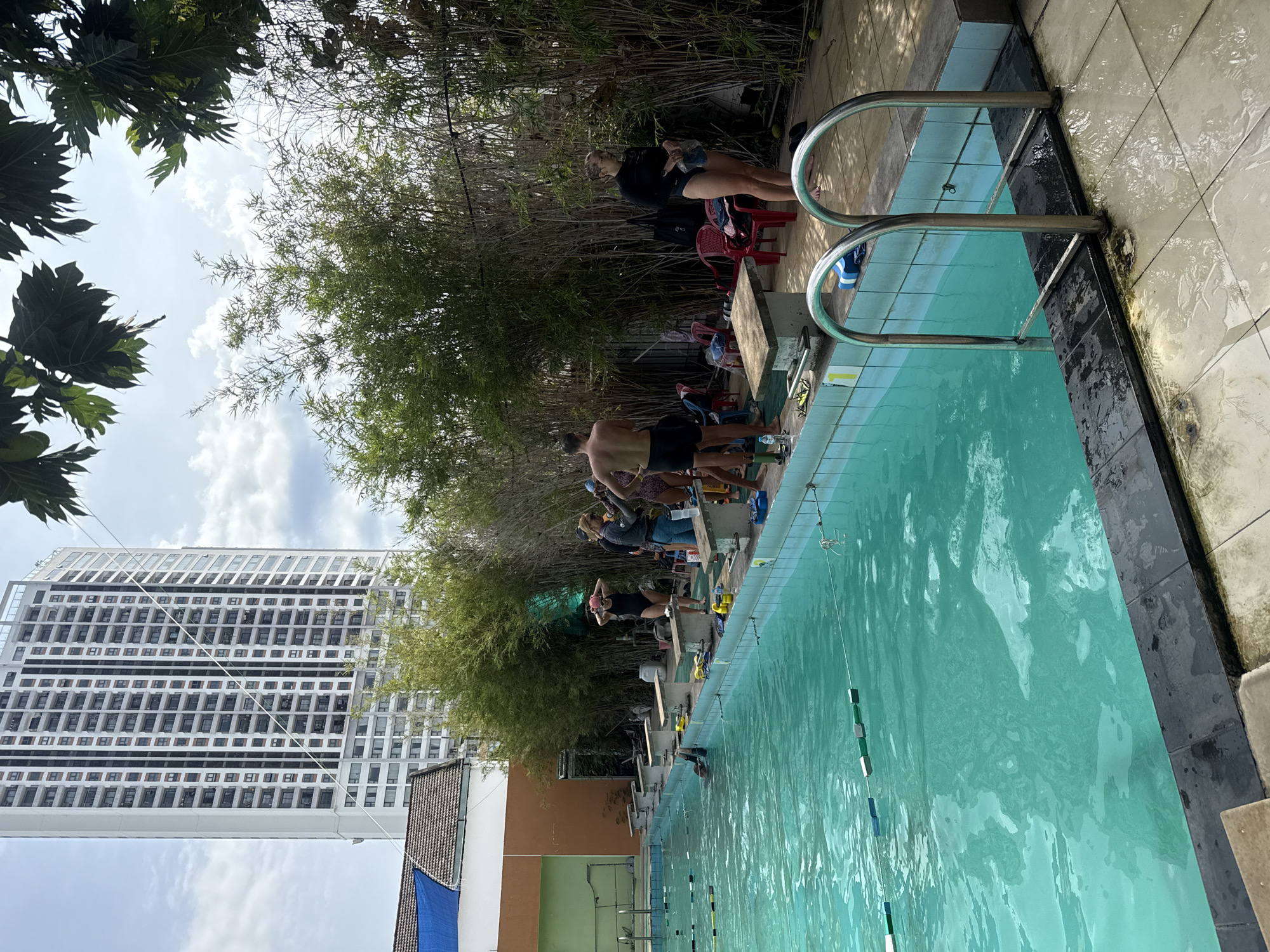 This image captures an outdoor swimming pool area likely located in Nha Trang Ward, Vietnam, given the architecture and vegetation. A tall, modern white building with many windows dominates the background, suggesting an urban setting. The swimming pool itself is rectangular with turquoise water and lane markers. Several people are visible around the pool, some in swimsuits and swim caps, appearing to be preparing for or having just finished swimming. There are diving blocks along the side of the pool, and bamboo plants line the perimeter of the area, providing a natural screen. Red plastic chairs are scattered around, and a tiled patio surrounds the pool. The overall impression is of a recreational facility in a tropical or subtropical climate.