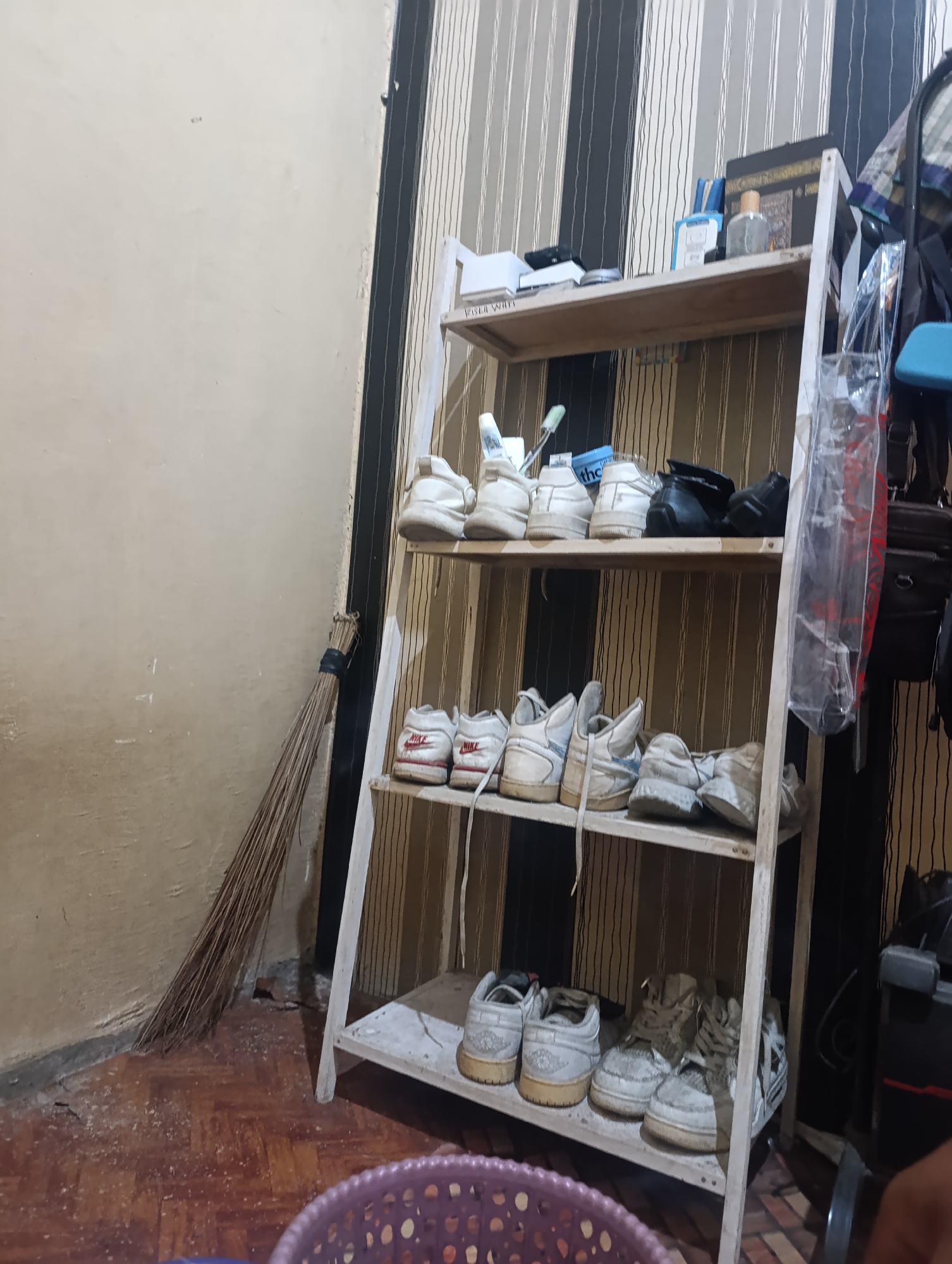 In Madatte, Indonesia, a narrow shoe rack stands against a wall adorned with black and beige vertical stripes. The rack is filled with various pairs of white sneakers, some appearing well-worn. On the top shelf, a few pairs of white shoes are visible, along with two black shoes. Below, the second shelf holds more white sneakers, including pairs with red Nike branding. The bottom shelf features a variety of white and grey sneakers, some of which are heavily scuffed. To the left of the shoe rack, leaning against the wall, is a broom made of natural fibers, its bristles tied together with black tape. The floor is a wooden parquet, scattered with light-colored debris. In the foreground, a portion of a purple plastic basket is visible. The scene appears to be an indoor storage area or hallway.