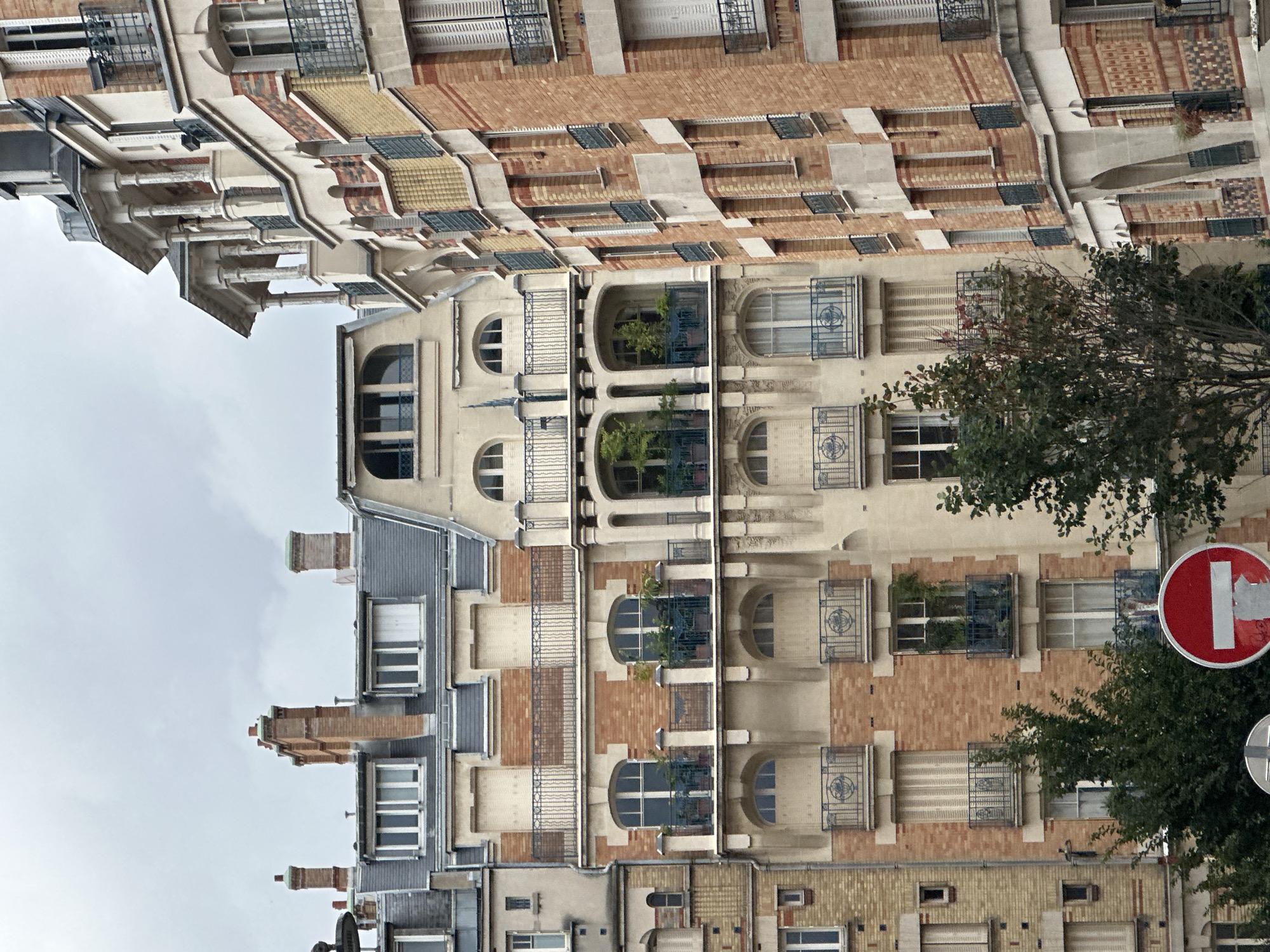 This is a medium-shot, eye-level, exterior, daytime, overcast photograph of a row of apartment buildings in Paris, France, likely in the 16th arrondissement given the architectural style. The buildings are tall and feature a mix of brick and stucco facades, with ornate balconies and numerous windows.
On the left, a lighter-colored building with a muted yellow and beige facade is partially visible. It has several rows of windows, some with shutters and some open. Chimneys with terracotta caps are visible on the roof.
The central building is predominantly beige with brick accents. It has multiple stories, each with balconies. The balconies are decorated with intricate wrought-iron railings and are lined with potted plants. The windows are arched on some levels.
The building on the right is an orange-red brick structure with decorative lighter-colored brick inlays. It also has balconies with wrought-iron railings and numerous windows, some with white shutters. A distinctive architectural feature on this building is a series of paired columns supporting a roofline detail.
In the foreground, to the left, a large tree with dark green foliage partially obscures the lower floors of the buildings. A red and white circular traffic sign with a horizontal white bar is visible in the lower left quadrant of the image. The sky is overcast and a pale gray.