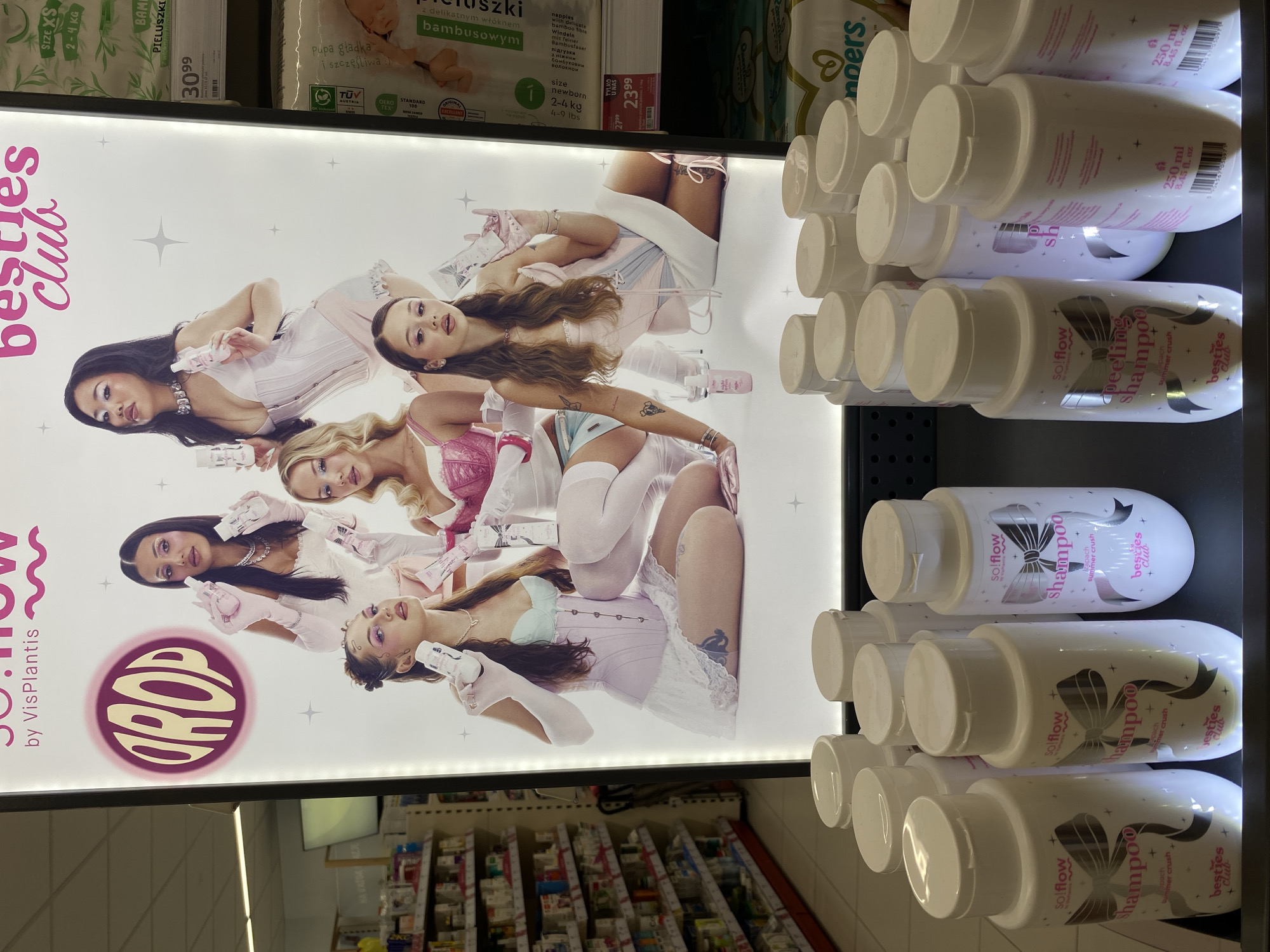The image shows a promotional display for "sol flow by VisPlantis besties club" in a store, possibly a pharmacy or supermarket, in Buk, Poland. The display features a large illuminated poster with five young women in fashionable outfits, holding bottles of product. Below the poster, a shelf displays several bottles of "sol flow shampoo" and "peeling shampoo." In the background, shelves stocked with various products are visible, along with a price tag indicating "23.99 zł." The lighting suggests an indoor retail environment.