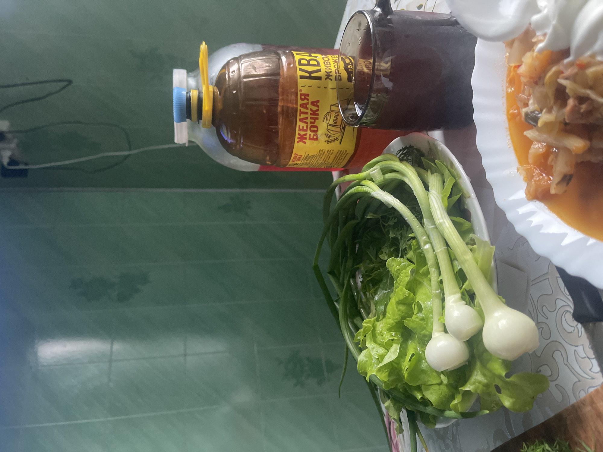 The image depicts a still life arrangement on a table, likely in a kitchen. In the background, a green tiled wall is visible, with some light reflections and what appears to be a shadow of a plant. In the foreground, to the left, a plate holds fresh green lettuce and several bulbs of green onions. To the right, a dark brown mug contains a dark liquid, possibly a beverage. Behind the mug, a large plastic bottle with a yellow label and a blue cap is prominent. The label is in Russian and includes the text "ЖЕЛТАЯ БОЧКА" (Yellow Barrel) and "КВАС" (Kvass). Another clear bottle with a red liquid is partially visible behind it. In the lower right corner, a white bowl contains a portion of what looks like soup or stew, topped with a dollop of white cream or sour cream. The overall scene suggests a meal is being prepared or has just been served. The location of Красное, Russia is mentioned in the prompt, but there are no visual cues in the image to confirm or deny this specific location.