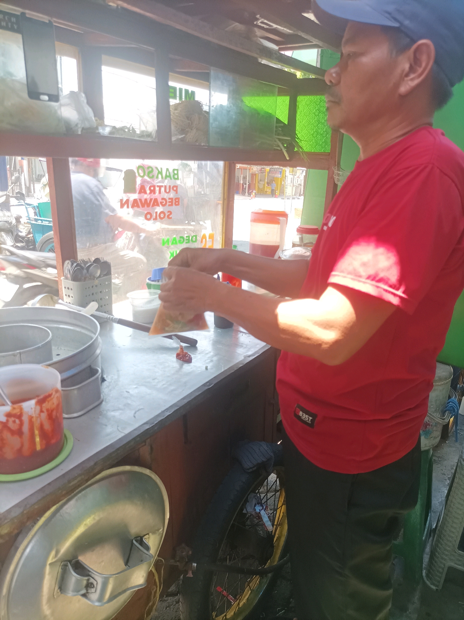 In Sumuragung, Indonesia, an outdoor food stall is depicted. A middle-aged man, wearing a red t-shirt and a dark blue cap, stands facing right, preparing food. He holds a plastic bag containing food in his hands, with his thumbs and index fingers pushing the contents down. In front of him is a metal counter. To his left, a metal container with a lid, possibly for food storage, rests on the counter. Further left, a large, cylindrical metal container with a perforated insert, likely for steaming or draining food, is prominent. A plastic tub filled with a reddish sauce is positioned in front of this container. Behind the man and to his left, a glimpse of a motorcycle and a blue cart is visible. The stall has a wooden frame with green translucent panels and a shelf above. On the shelf, a mobile phone is propped up, and bundles of dried straw or noodles are stored. Behind the counter, there's a glimpse of a street scene with buildings and people. The overall environment suggests a busy, everyday market or street food setting.