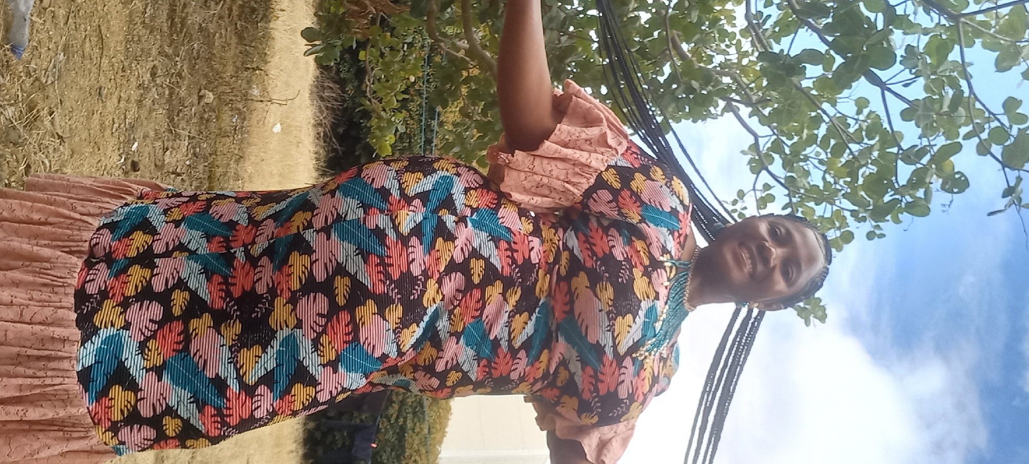 A full-body shot captures a Black woman standing outdoors, possibly in a residential area of Jos, Nigeria, given the visible vegetation, dry grass, and a building in the background. The woman is smiling at the camera, and her long, braided hair extends behind her. She is wearing a colorful, knee-length dress with a tropical leaf print in shades of pink, yellow, teal, and white against a dark background. The dress features ruffled sleeves and a ruffled hem, and she is accessorized with a beaded necklace. The environment includes a large tree with green leaves to her left, telephone wires overhead, and a dry, grassy ground with some scattered debris. The lighting suggests it is daytime.