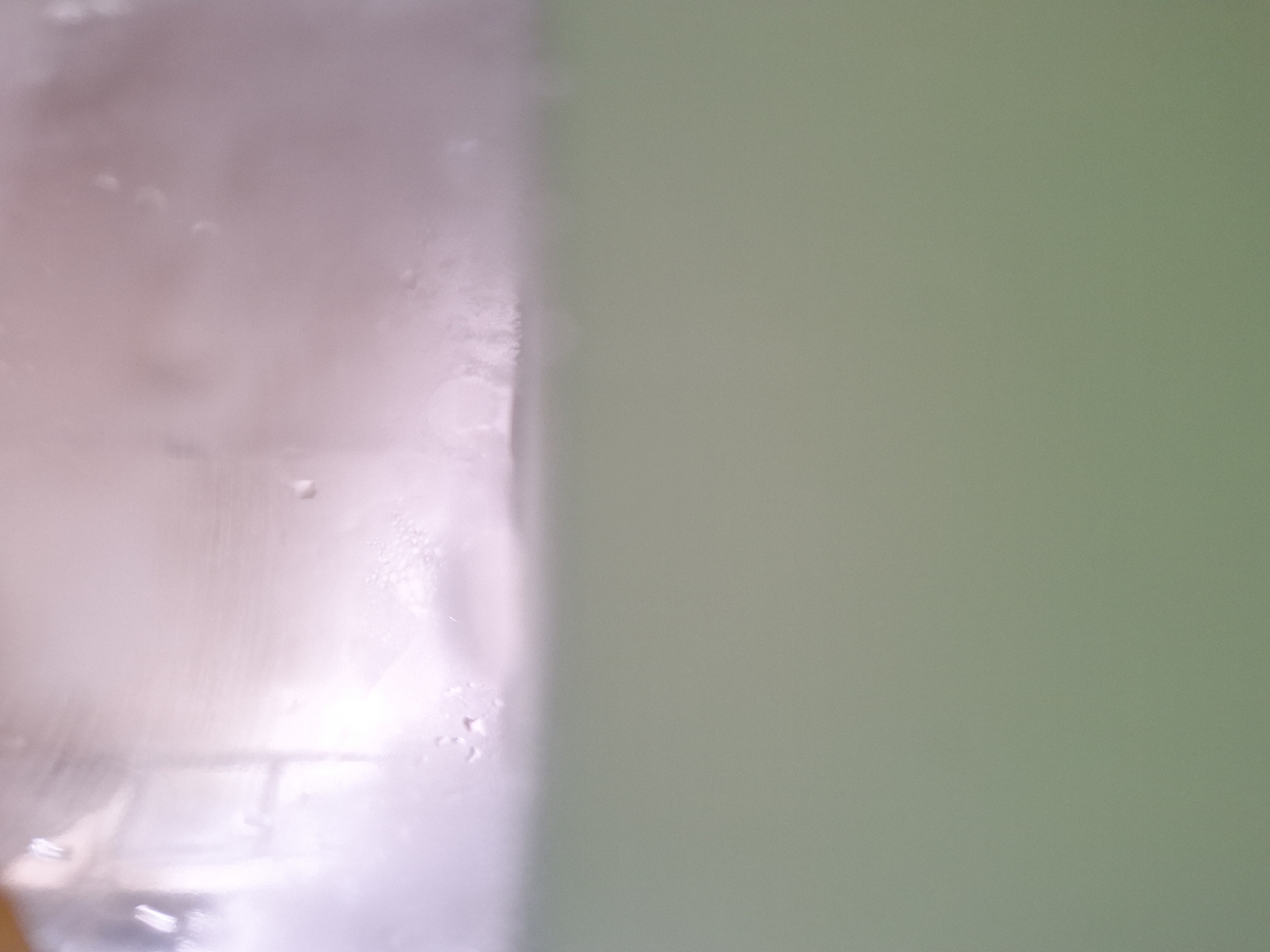 This image captures the internal view of a transparent container holding an opaque, muted green liquid. The upper approximately one-third of the frame displays the container's interior surface, which exhibits soft, blurry reflections of ambient light sources, predominantly in white and faint pinkish-purple tones. Numerous small water droplets are visible on this surface, indicating condensation. A distinct, slightly wavy horizontal line marks the interface between the container's interior and the liquid, with minor textural irregularities or small bubbles visible at the liquid's surface, particularly towards the right. The lower two-thirds of the image consist entirely of the uniform, solid muted green liquid, which appears completely opaque and devoid of any internal features. No individuals, specific actions, or direct interactions are depicted. The environment is inferred to be indoors, under ambient lighting conditions, and the scene is located in Casablanca, Morocco.