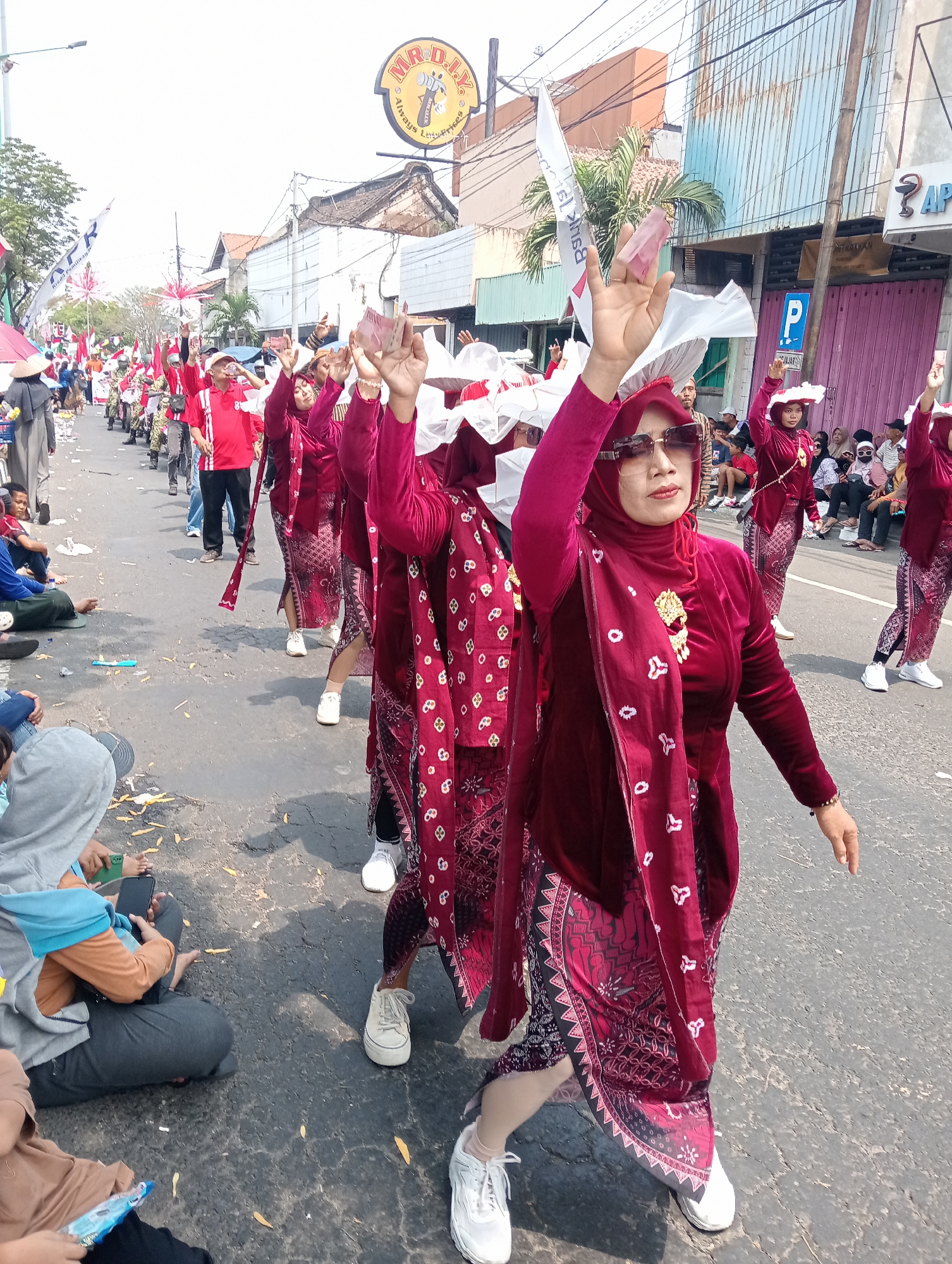 In Demak, Indonesia, a parade is underway on a street lined with buildings and businesses. The foreground features several women in traditional maroon attire, some with white head coverings, dancing or posing. One woman in the front right wears sunglasses and a gold brooch. They are holding what appear to be banknotes in the air. In the background, a procession of people in red shirts and others in traditional clothing are visible, along with spectators sitting on the side of the road. A sign for "MR.D.I.Y" is prominent on a building. The scene suggests a festive or celebratory event.