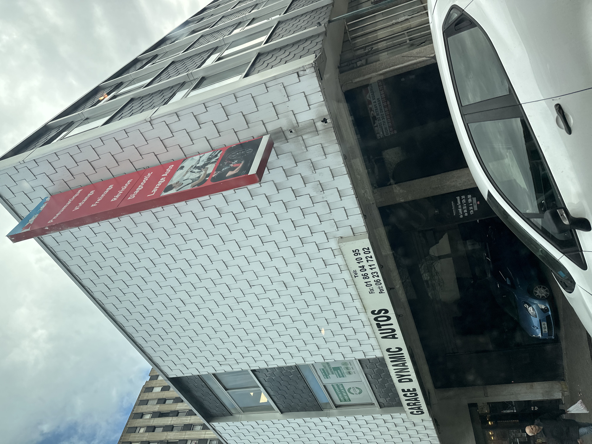 The image shows a multi-story building with a car garage named "GARAGE DYNAMIC AUTOS". The building's exterior is covered with white and grey tiles. A red sign on the building lists various car services in French, including "Pneumatique, Vidange, Freinage, Révision, Diagnostic, Lavage Auto". A blue car with the license plate "GP-908-HV" is parked in the garage. A white car is visible on the right side of the image. Below the "GARAGE DYNAMIC AUTOS" sign, there is another sign, which says "Tél, Fix: 01 86 04 10 95, Port: 06 23 11 72 02". The sign "CENTRE D'EXAME CODE DE LA ROUT LE CODE AVEC OU SANS RESULTAT INSTANTANE REPASSEZ EXAMEN DIRECTEMENT" is visible near a window on the building. A man is seen standing near the entrance of the garage. The location is likely Sarcelles, France, as the phone number code "01" is associated with the Paris region. Another taller building is visible in the background. The sky is cloudy.