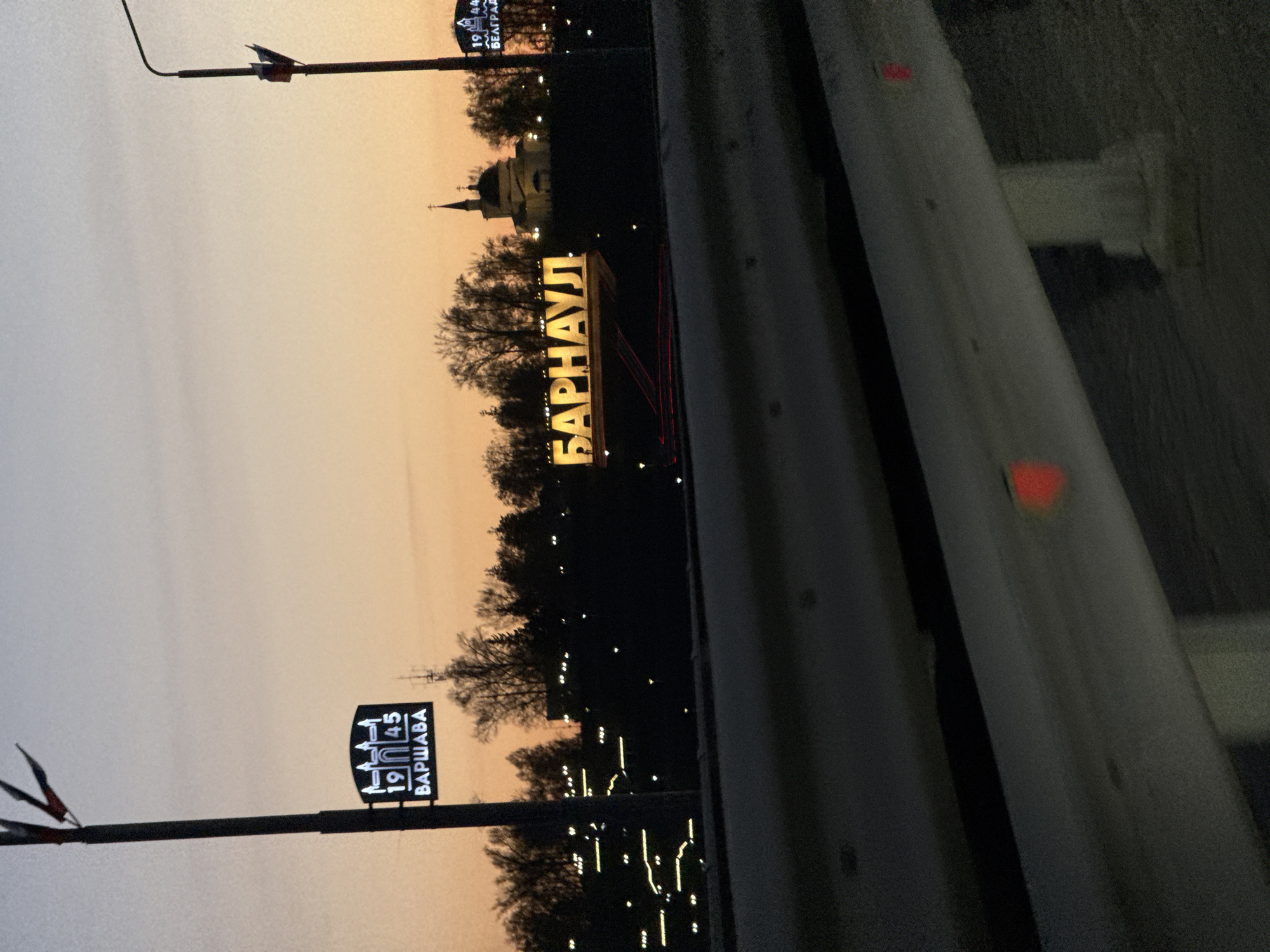 The image captures an urban scene at twilight or dusk, likely viewed from a vehicle on a road or bridge. The sky transitions from a warm orange near the horizon to a muted grey-blue overhead.
The most prominent feature is a large, illuminated sign spelling "БАРНАУЛ" (Barnaul) in yellow Cyrillic letters, situated in the midground. To the left, two smaller, illuminated signs on poles display text: one shows "1944" and "БЕЛГРАД" (Belgrade), while the other indicates "1945" and "ВАРШАВА" (Warsaw). These likely serve as commemorative or historical markers.
In the background, silhouetted trees line the horizon, interspersed with faint, distant city lights. A distinctive building with a steeple and dome, possibly a church or historical structure, is visible in the distance, partially illuminated.
On the right side of the frame, a grey guardrail or barrier runs diagonally, featuring small red reflective elements, suggesting the viewpoint is from a roadway or an elevated structure. No specific activity or people are visible, presenting a static, serene view of the cityscape at the golden hour. The signs clearly identify the location as Barnaul.