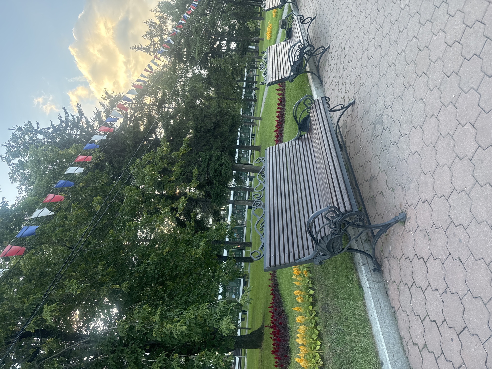 The image depicts a park scene in Irkutsk, Russia, featuring several park benches along a paved walkway. The benches are made of dark wood slats with ornate black metal legs and armrests. Behind the benches, a manicured lawn is visible, with flowerbeds featuring red and yellow blooms. A line of trees, primarily deciduous and coniferous, forms a backdrop, with some of the trees showing signs of autumn foliage. Above the trees, a string of colorful flags (red, white, and blue) is strung across, suggesting a festive occasion or national celebration. The sky is partly cloudy with the sun breaking through, casting a warm light. In the distance, behind the trees and lawn, a long, dark structure, possibly a train or a building, is partially visible. The paved walkway is made of interlocking, light-colored bricks arranged in a geometric pattern. The overall impression is one of a tranquil public space, well-maintained and decorated for an event.