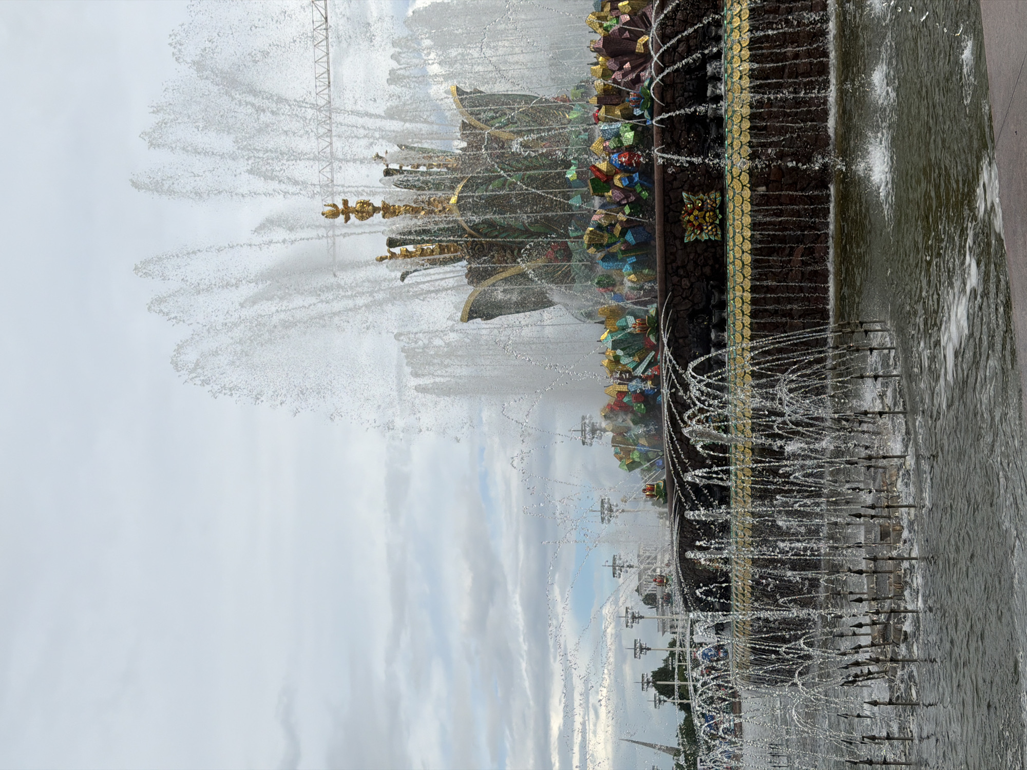 The image shows a fragment of the Friendship of Peoples Fountain in Moscow, Russia. The fountain is elaborately decorated with gold and colorful ornaments. Water jets spray upwards and outwards from various points. The background is a cloudy sky. The fountain is located in a public park.