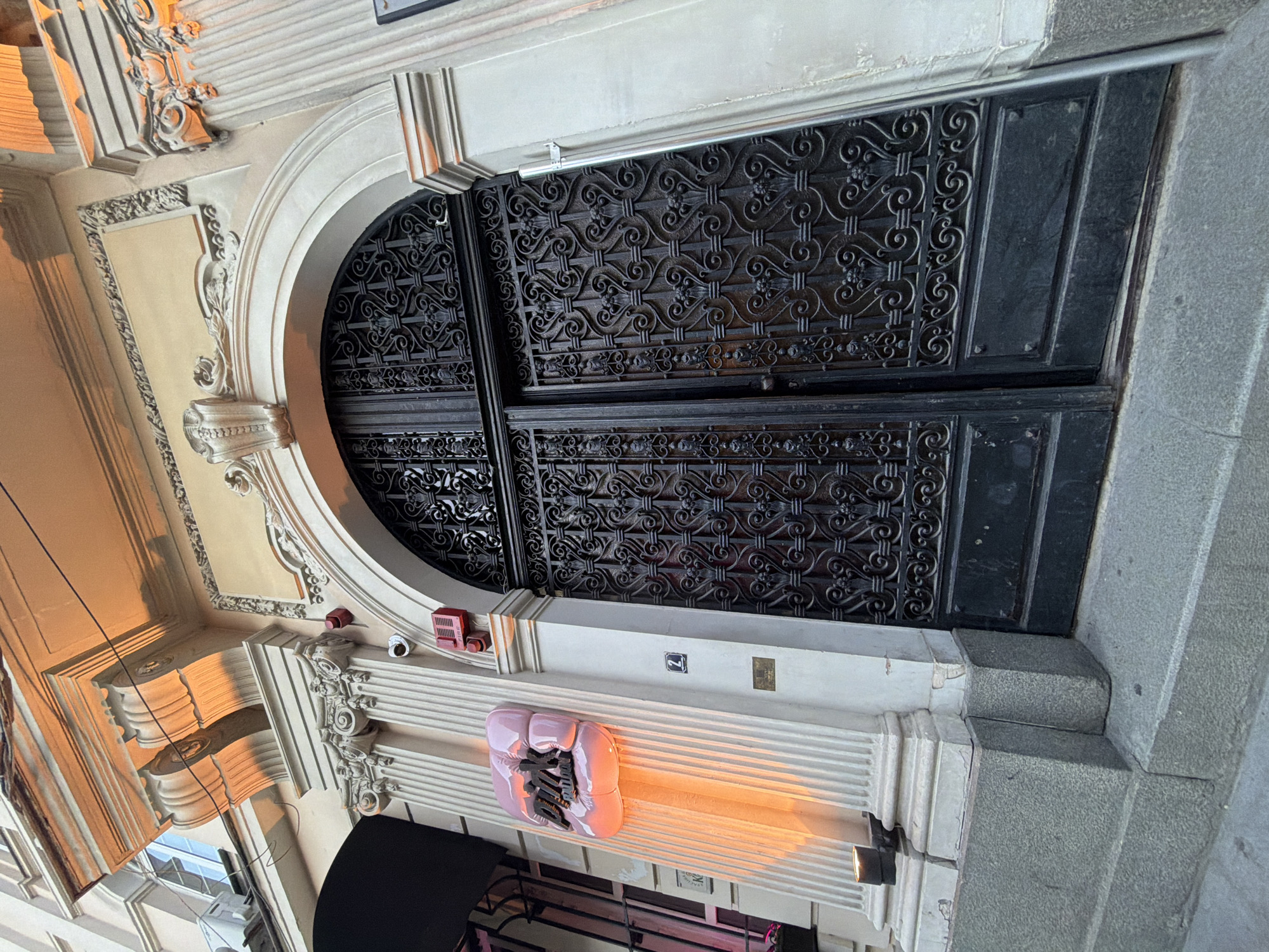 This image captures the ornate facade of a building, focusing on an arched doorway with a decorative black metal gate. The architecture features classical elements like columns and intricate moldings.
The subjects include an abstract pink art piece mounted on the facade, illuminated by warm, orange light that suggests it is either dusk or artificial lighting. There are also some small, red fire alarm devices visible. The scene is an exterior, likely a street or building entrance, with the lighting indicating a time of day when artificial lights are active.
Notable details include the elaborate scrollwork on the metal gate, the layered stonework and plaster details of the building's exterior, and the soft glow emanating from the art piece. The image is taken from a low angle, looking upwards towards the doorway.
Visible text includes the number "2" on a small square plaque on the wall above the art piece.