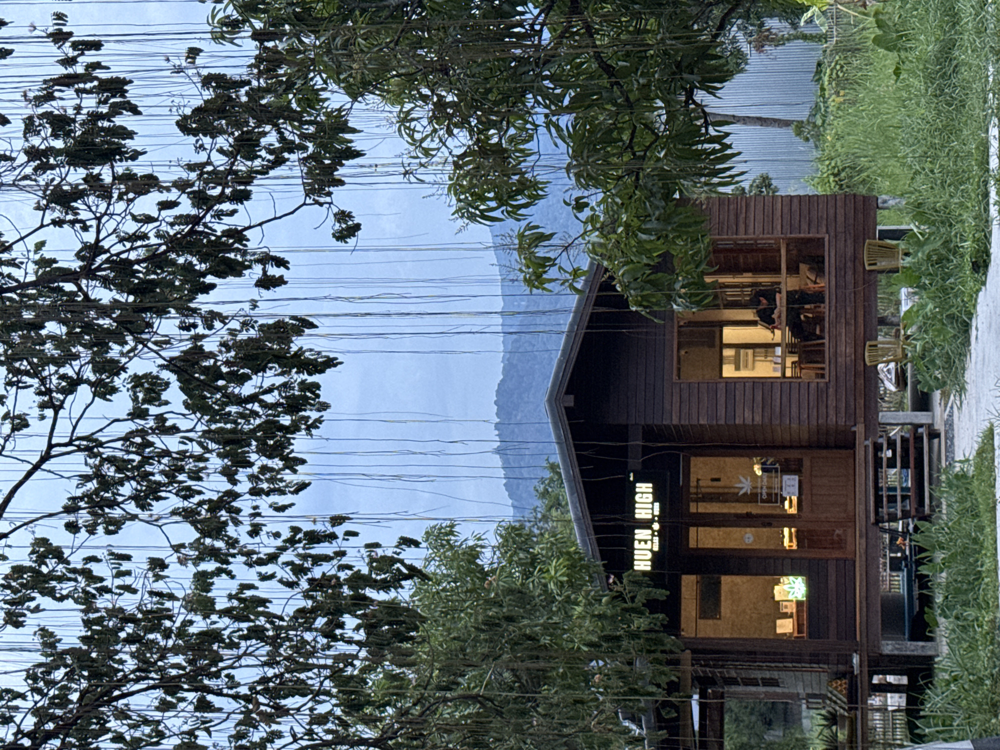 The image shows a wooden building, likely a shop or cafe, with the name "Huen High" displayed vertically. The building has large windows revealing interior lighting and silhouettes of people inside. Trees flank the building, partially obscuring the view. Power lines stretch across the sky. In the background, a mountain range is visible. The location is Chiang Mai City Municipality, Thailand.