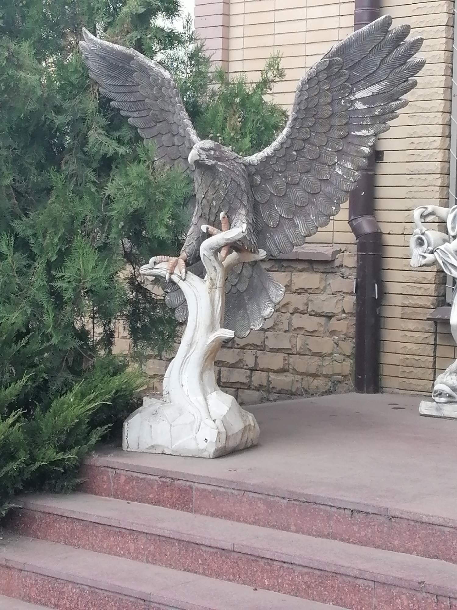 This is a full outdoor shot of a statue of an eagle with its wings spread wide, standing on a white, stylized tree stump. The statue is made of a dark, textured material, possibly bronze or concrete. The eagle's head is turned to the left, and its talons are gripping the stump. The eagle statue is positioned on a concrete platform in front of a brick building. To the left of the eagle, there is a dense green shrub. To the right of the eagle, a dark downspout runs along the side of the building. Further to the right, partially visible, is another statue, appearing to be a classical figure made of white material. The foreground shows a set of granite steps leading up to the platform. The lighting suggests it is daytime. The location is identified as Rostov, Russia.