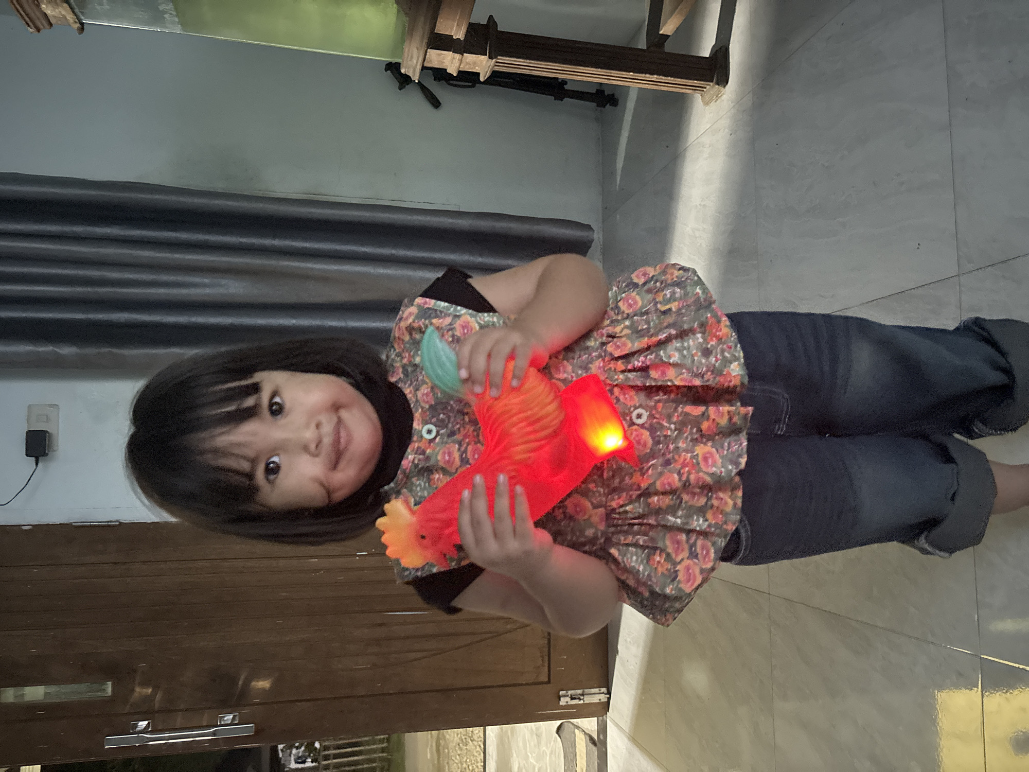 A young girl with dark hair and bangs stands in what appears to be a home or indoor setting. She is holding a bright red, illuminated toy rooster in both hands. The toy has yellow and green accents. The girl is wearing a dark top with a floral-patterned vest over it, and dark denim shorts. The flooring is tiled with a light gray pattern. In the background, a wooden door is visible to the left, and grey curtains hang to the right. A table with a tall, green glass object is also present on the right side of the frame. The environment suggests a domestic setting within the city of Lhokseumawe, Indonesia.