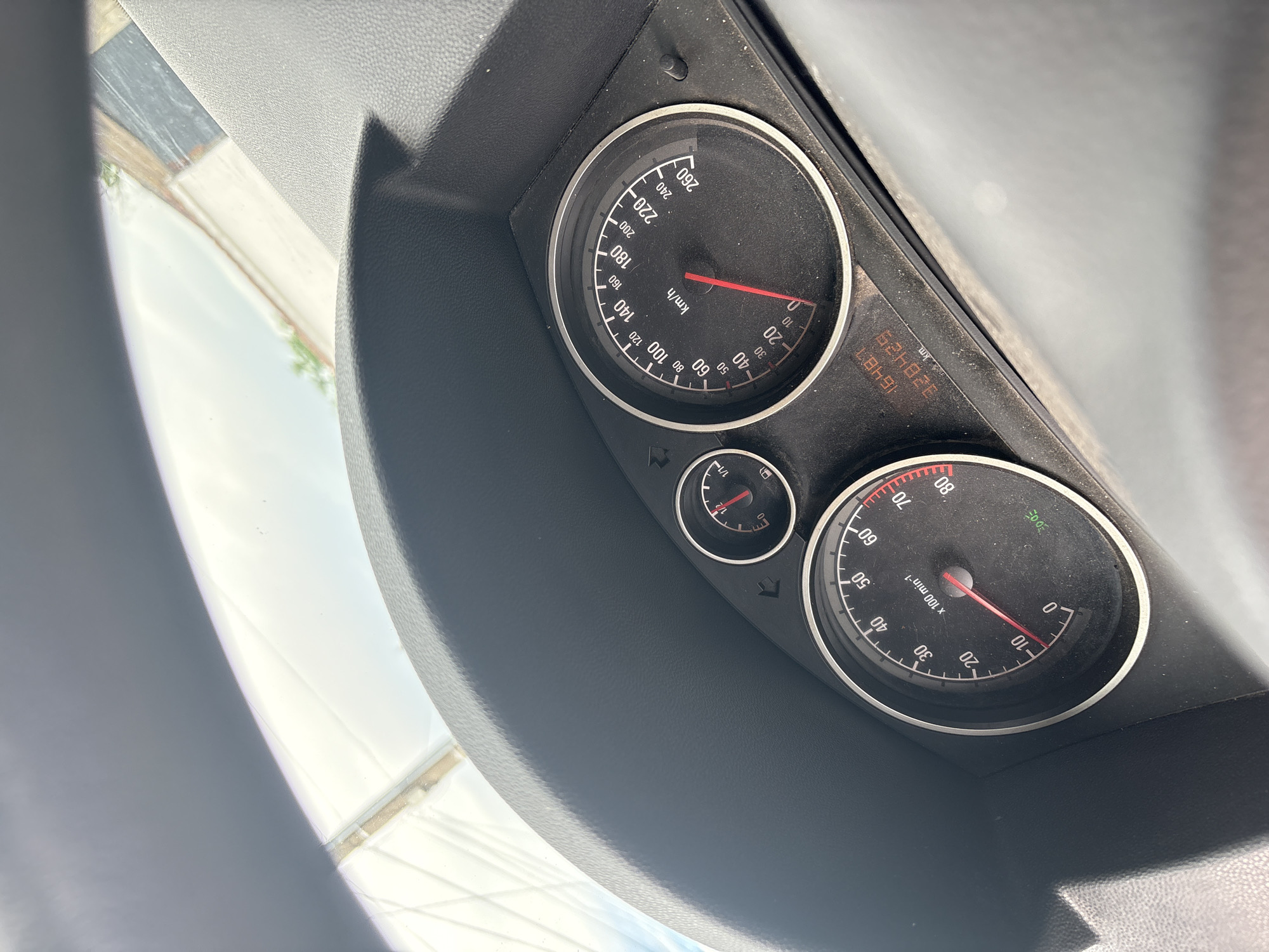The media file displays the dashboard of a car, focusing on the instrument cluster. The scene is illuminated by bright daylight, with a clear sky and faint outlines of structures visible through the windshield. The dashboard itself is dark grey and shows some dust accumulation.
The main subjects are the car's gauges. A prominent speedometer, marked "km/h", shows a reading of 0 km/h, indicating the vehicle is stationary or moving very slowly. Its dial ranges from 0 to 260 km/h. To its right, a digital display shows the total odometer reading of "328429 km" and a trip meter reading of "16487". Below the speedometer, a smaller fuel gauge is visible, with markings "0", "1/2", and "1/1".
To the right of the fuel gauge, the tachometer is present, marked "x 100 min-1" and scaled from 0 to 80. Its needle also rests near 0. Several small indicator lights are visible around the gauges, including a green icon that resembles a headlight or parking light symbol. A portion of the steering wheel is visible on the left side of the frame. The overall setting is inside a car located in Karadjovo, Bulgaria.