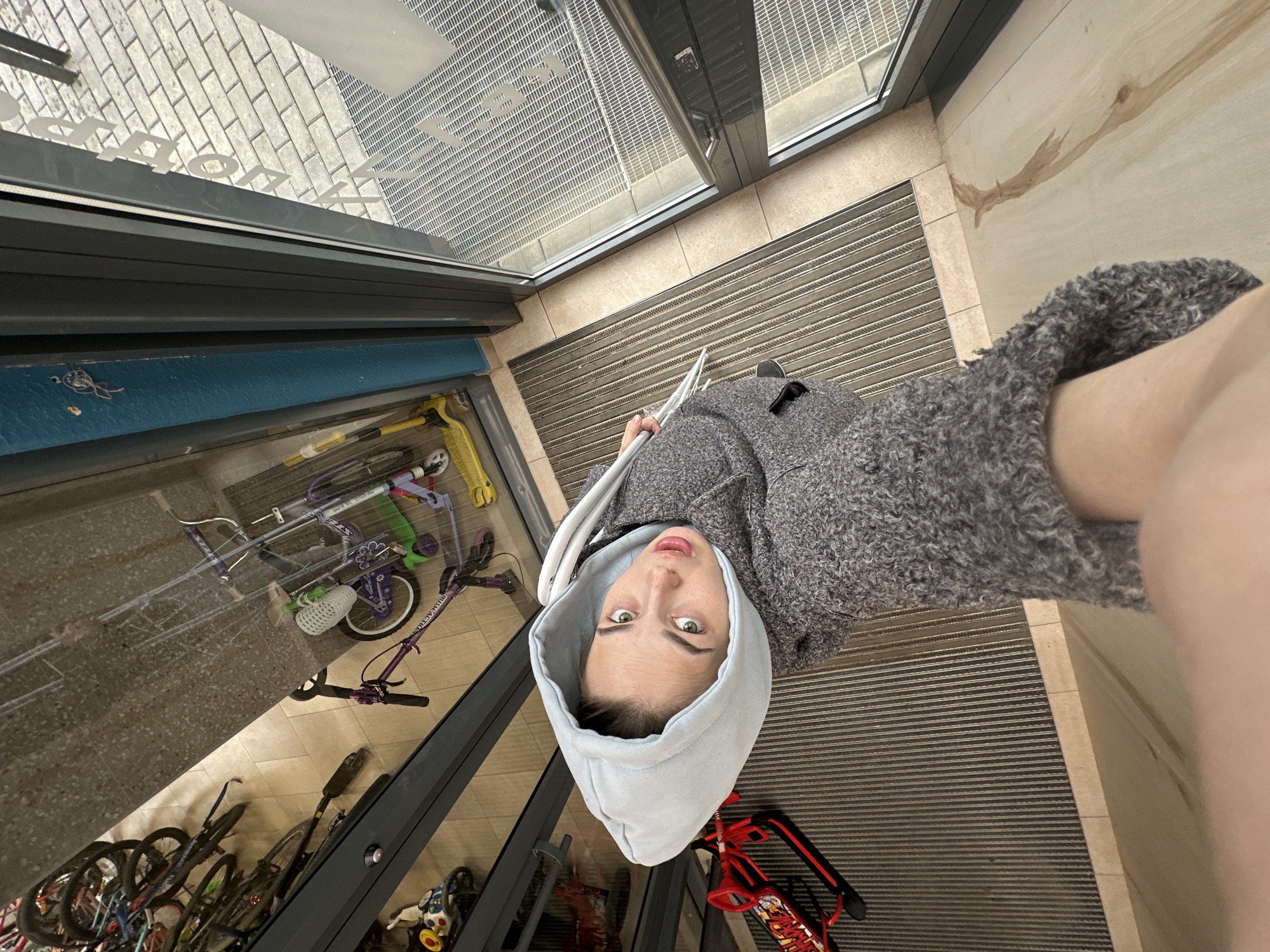 A woman wearing a gray hooded sweatshirt and a gray wool coat is looking at the camera. The image is taken from above, appearing as if the woman is looking up at the camera.
The environment appears to be the entrance of a building, possibly an apartment complex. Inside the glass door, a collection of bicycles and scooters are visible, suggesting a designated storage area. The flooring immediately inside the entrance is a gray, ridged mat. Outside the glass door, there's brick-patterned paving.
Given the city (Perm, Russia) specified in the prompt, the location could be a residential building's entrance with a designated bike and scooter storage area.