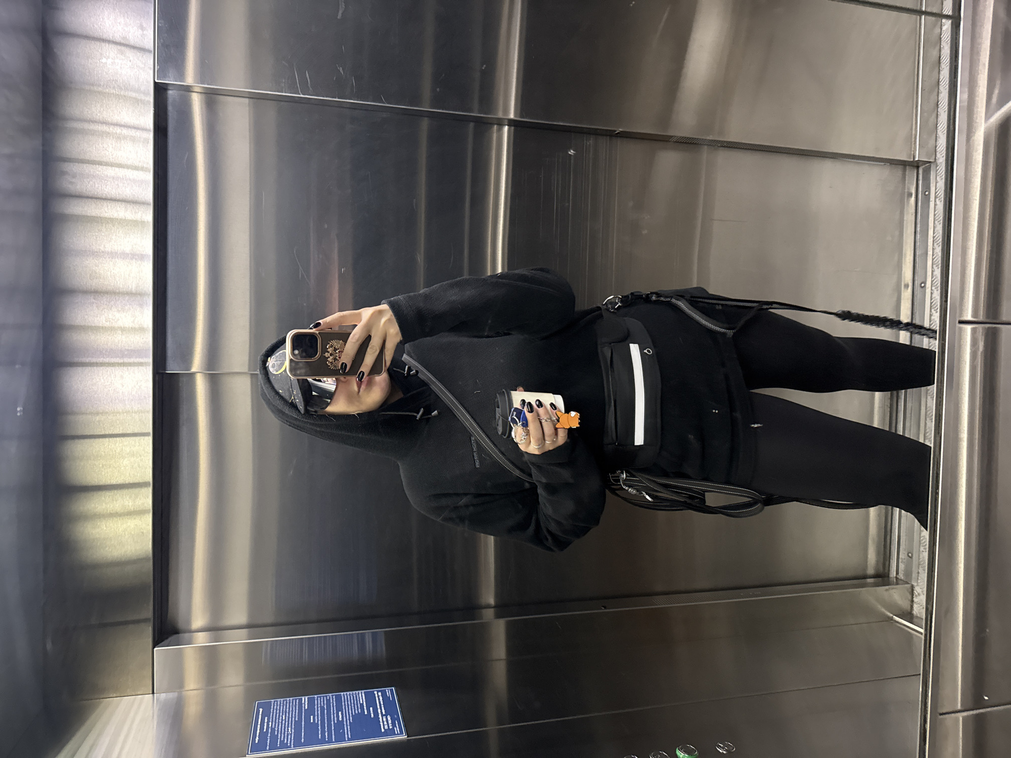 A person in black clothing takes a mirror selfie in a stainless steel elevator. They are holding a phone in their right hand and an object (possibly a vape pen) in their left. The elevator appears to be inside a building in Krasnoyarsk, Russia, as suggested by the language on the informational sign located inside the elevator.